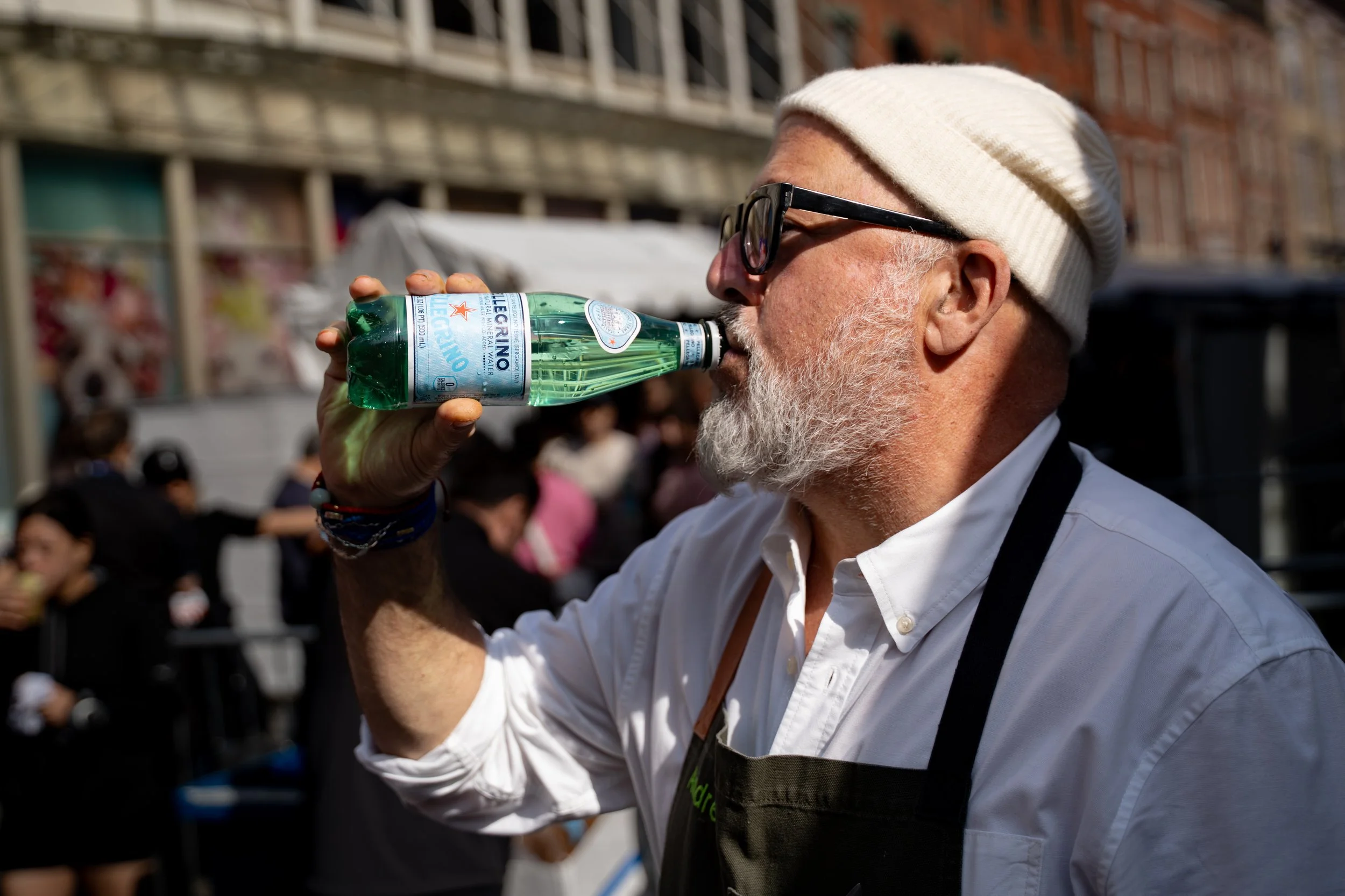 An older man with a white beard and glasses wearing a beige knit hat and a white shirt is drinking water from a green Heineken bottle in an outdoor urban area with people and buildings in the background.