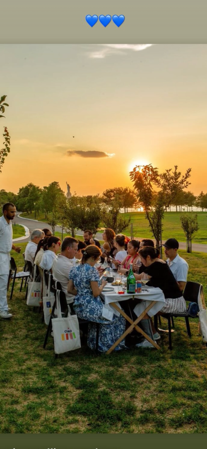 People dining outdoors at a long table during sunset, with a park and the Statue of Liberty in the background, and blue hearts at the top of the image.