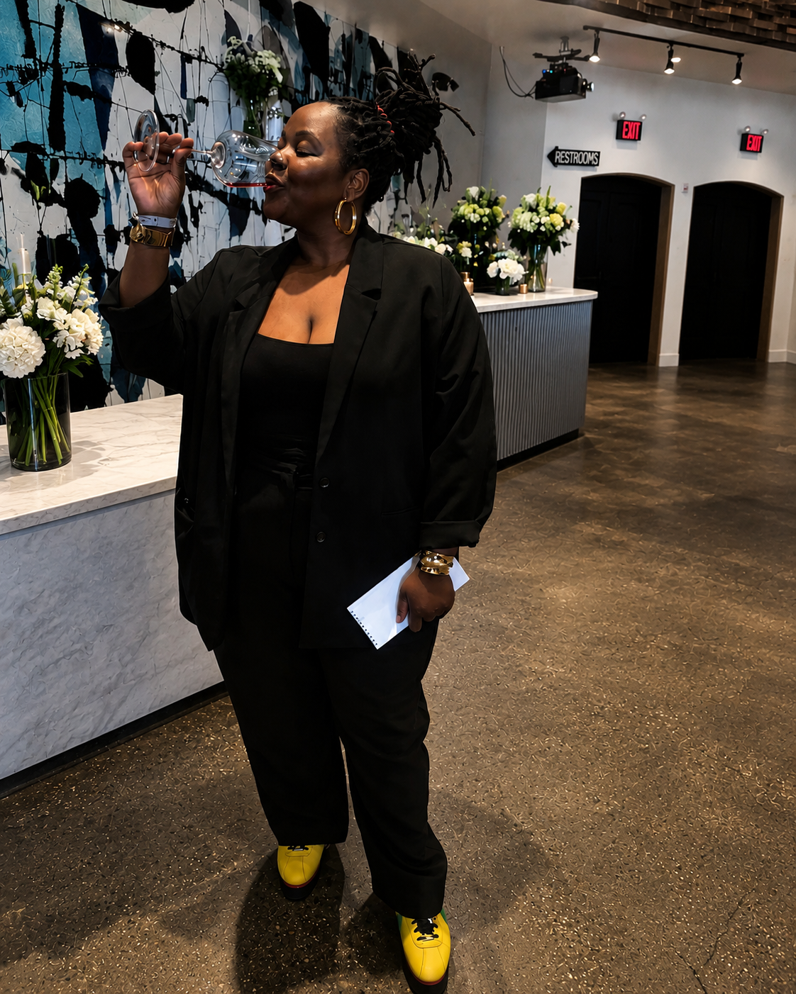 Woman with dreadlocks drinking from a glass at an indoor event, with floral arrangements and abstract wall art in the background.