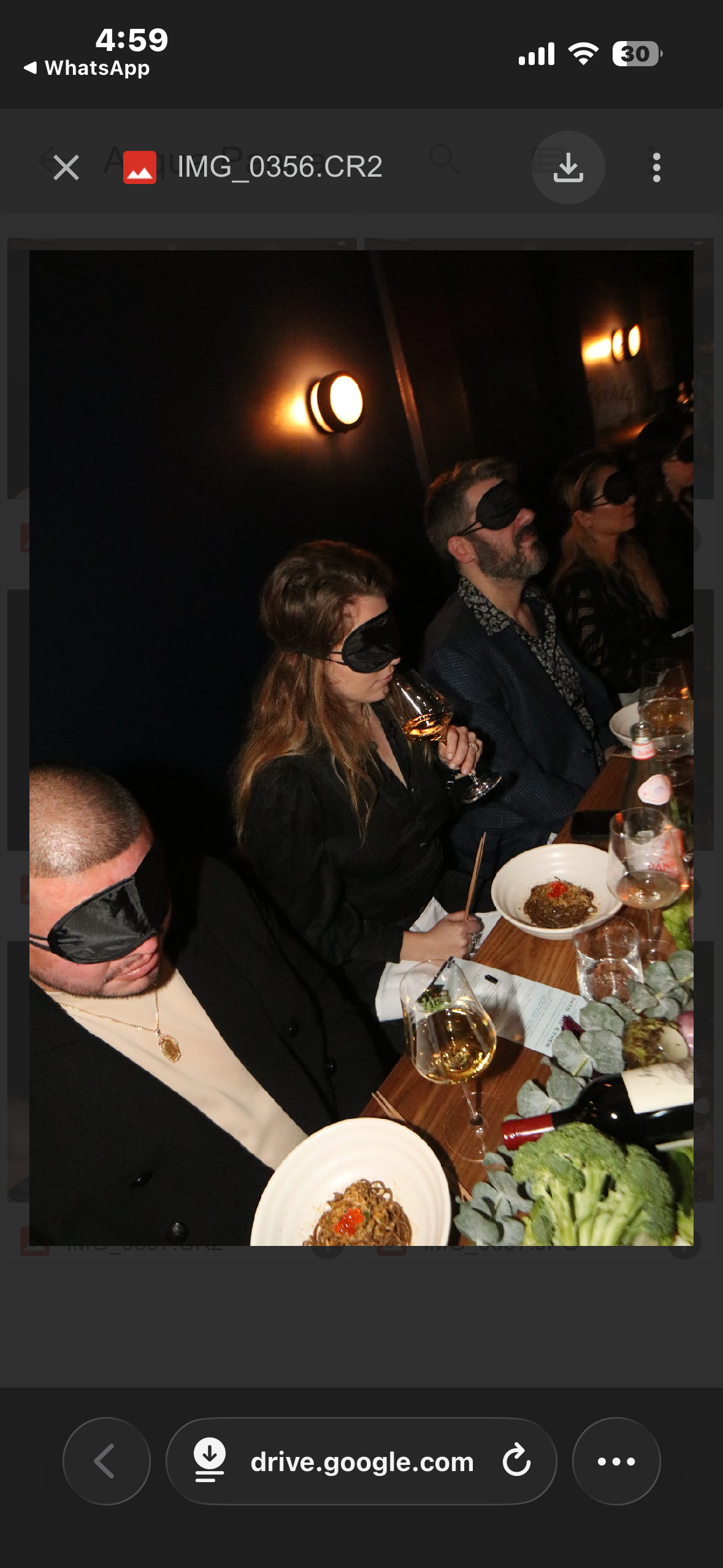 People sitting at a dinner table wearing black blindfolds, with drinks and plates of food, in a dimly lit room.