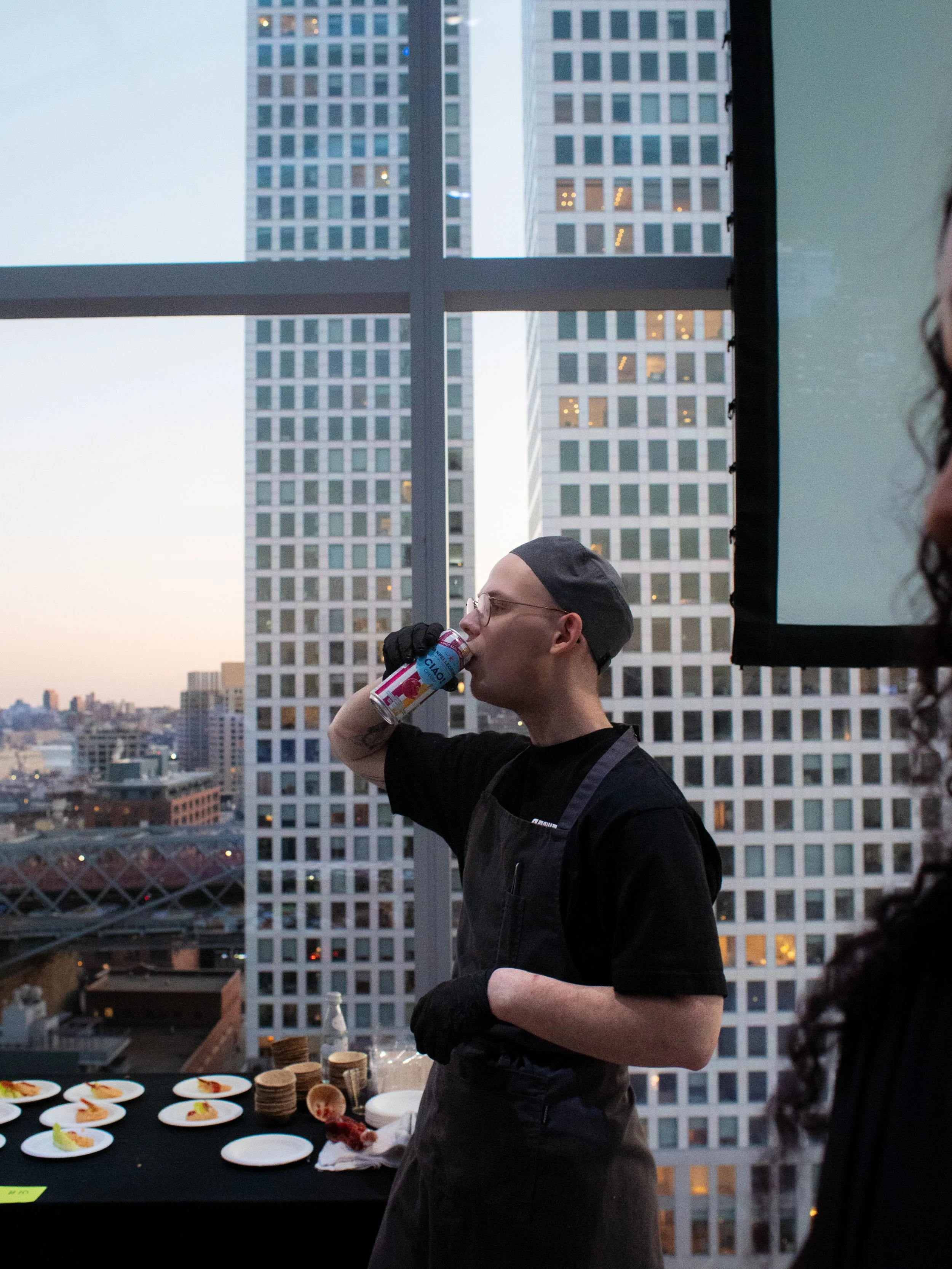 A person wearing glasses, a black hair cap, and black gloves is drinking from a can on a balcony in an urban setting with tall office buildings in the background. There is a table with small plates of food in front of them.