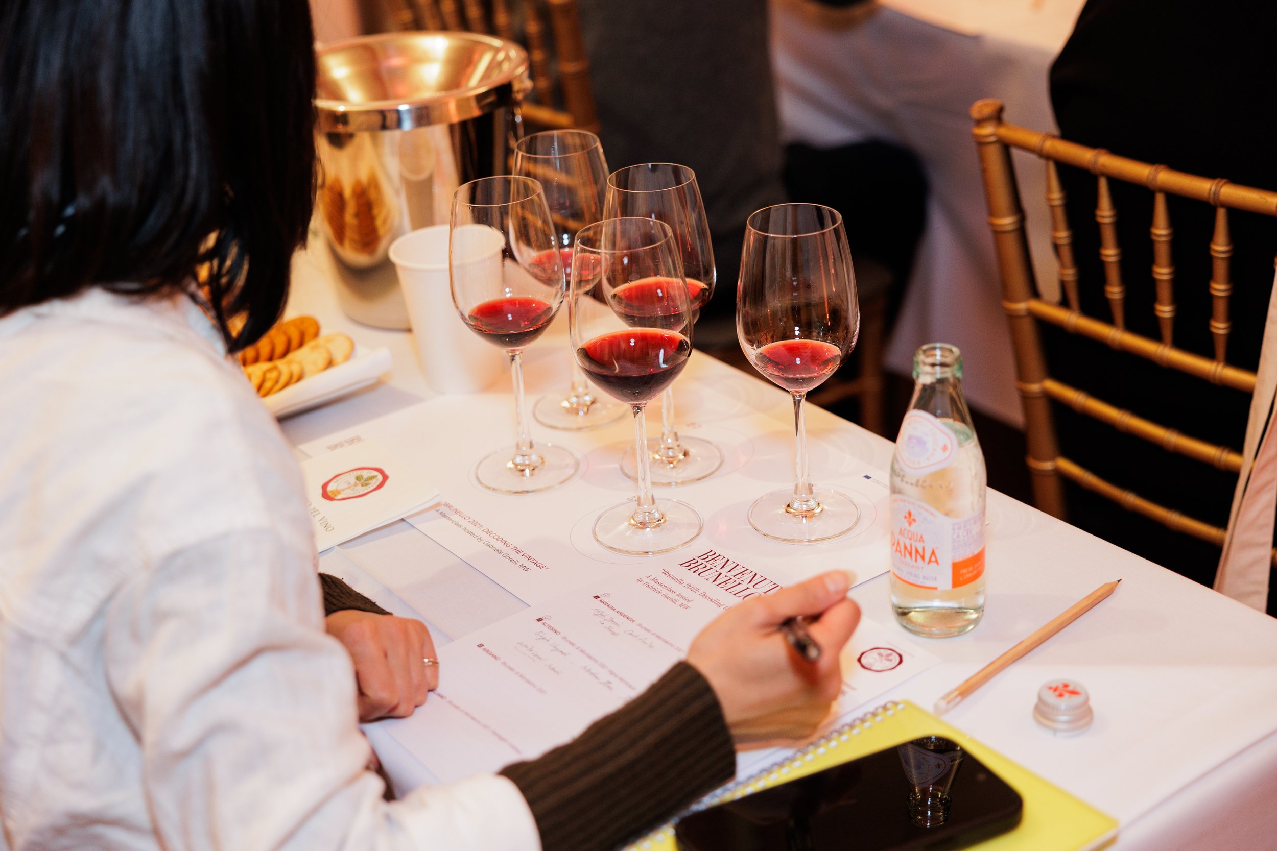 A person wearing a white sweater with brown and black sleeves is sitting at a table with four glasses of red wine, a menu, a pencil, and a clear bottle of water. There is a metallic ice bucket and snacks in the background, and other people are seated around the table.