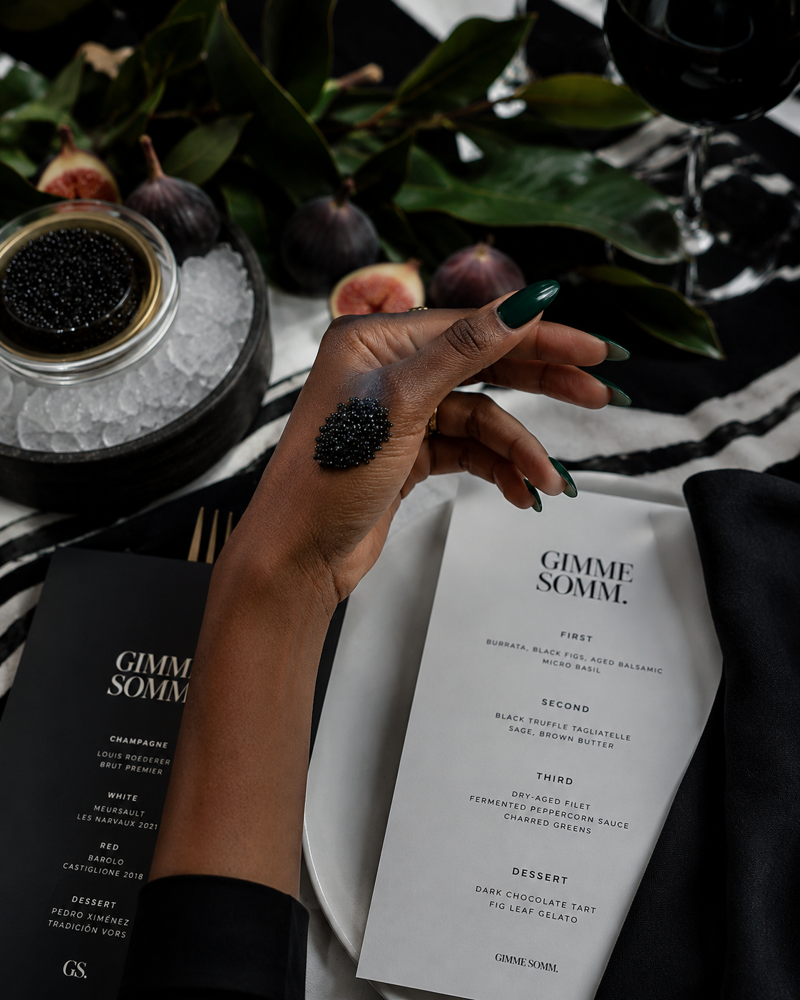 Close-up of person's hand with green nails, adorned with black caviar on finger, holding a black caviar spoon. In the background, a white menu with black text lists a multi-course meal, with figs and a glass of red wine nearby.