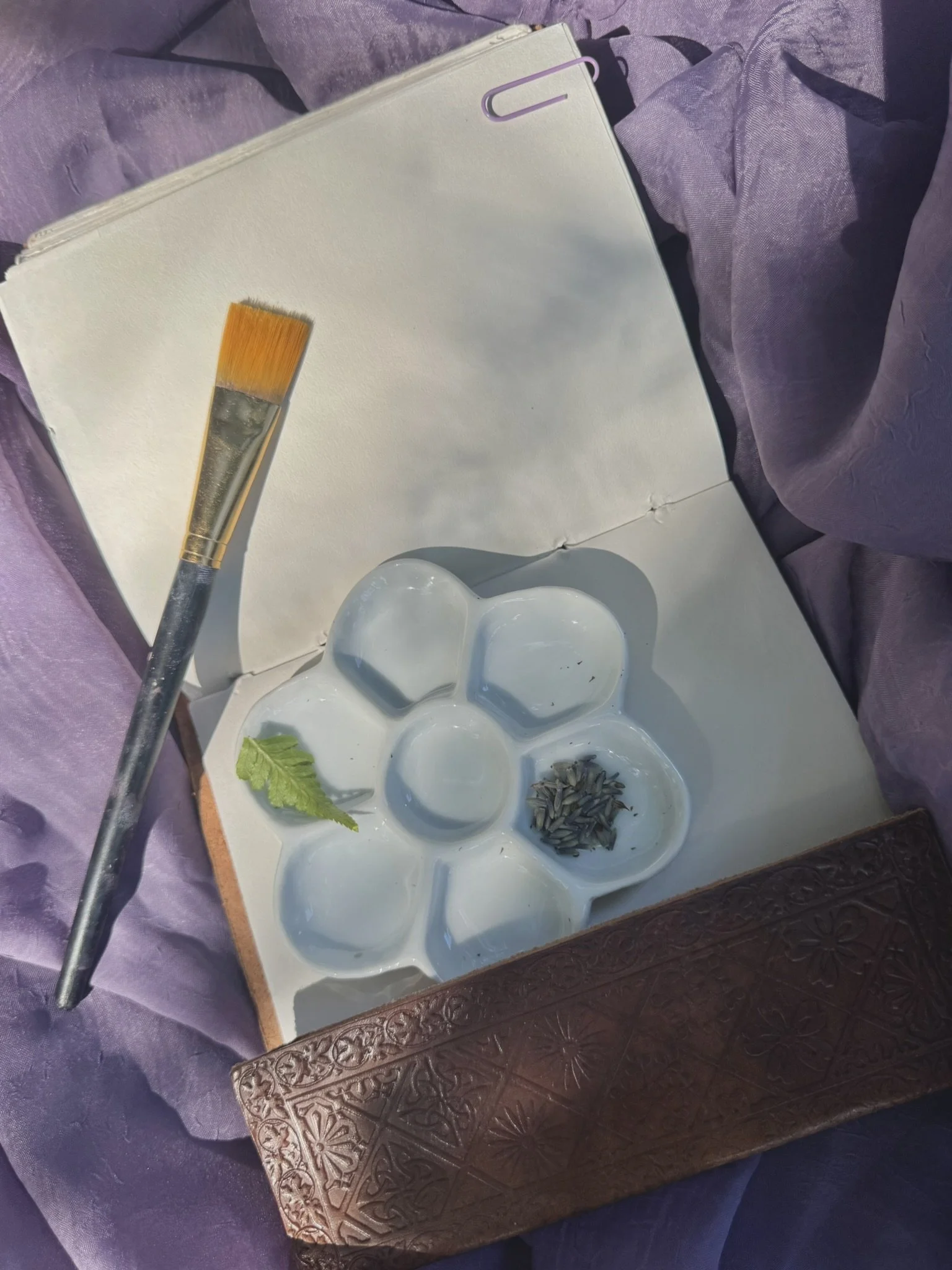 A paintbrush resting on a white palette with small compartments containing seeds, a green leaf, and white paint. The palette is placed on a decorative wooden surface, with purple fabric around.