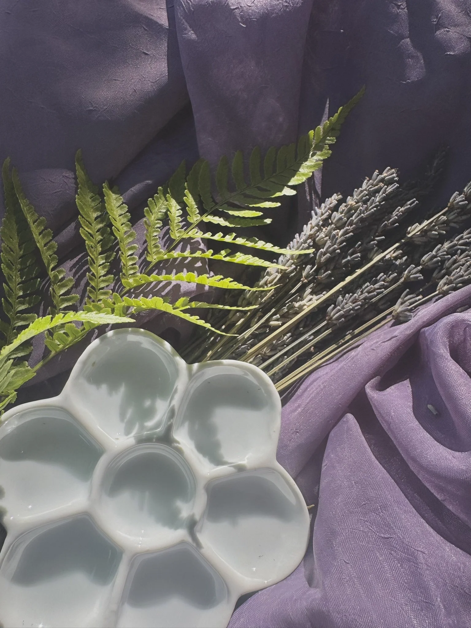 A purple cloth with some torn and distressed areas, a green fern leaf, dried lavender, and a white flower-shaped ceramic dish with water.