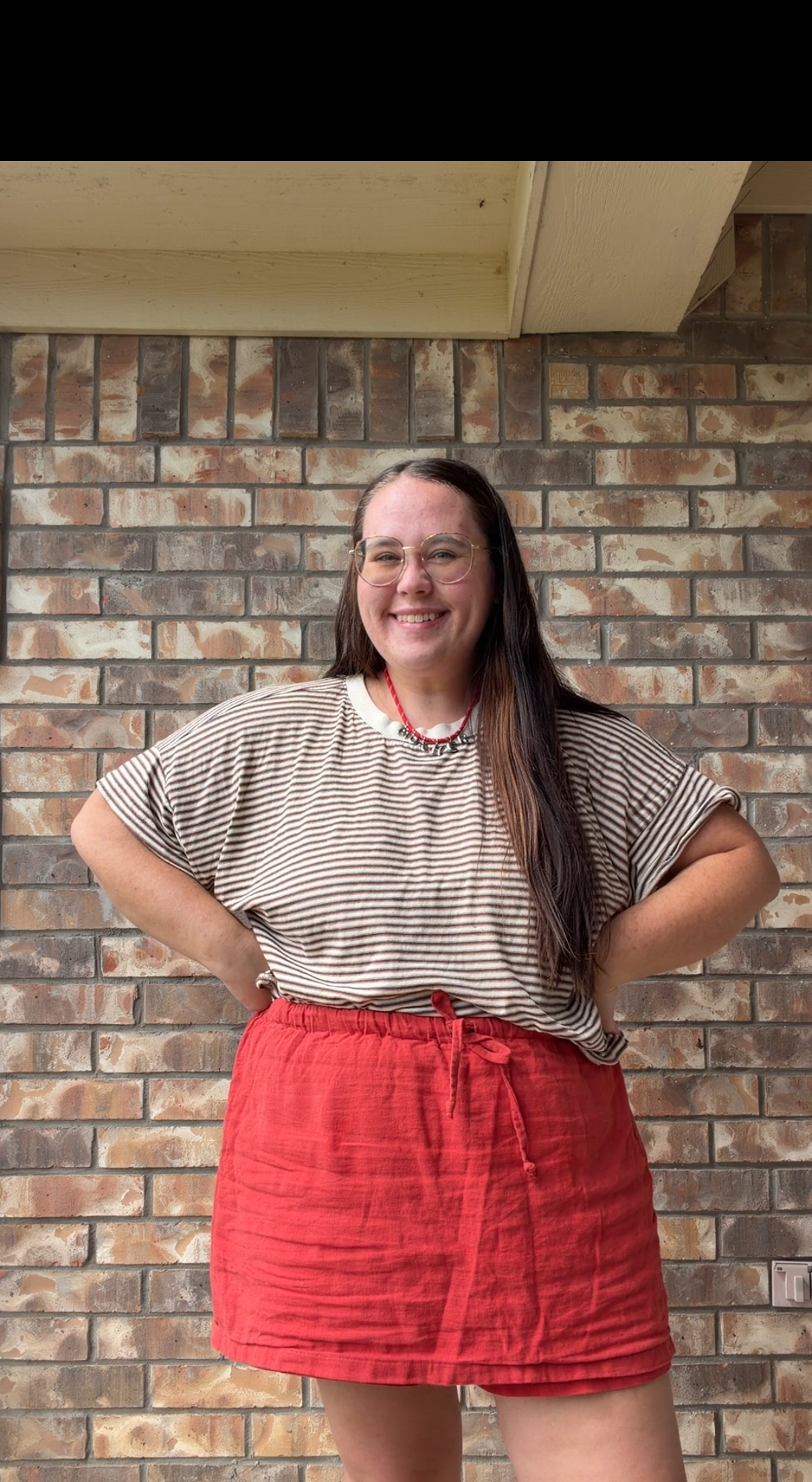 A woman with glasses and long dark hair stands in front of a brick wall, smiling with her hands on her hips. She is wearing a striped beige shirt and a red skirt.