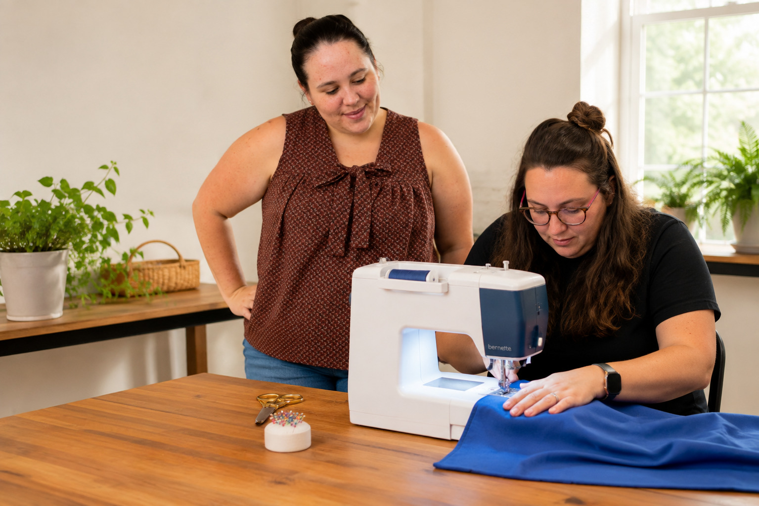 Two women working with a sewing machine; one is sitting and sewing a piece of blue fabric, while the other stands observing.