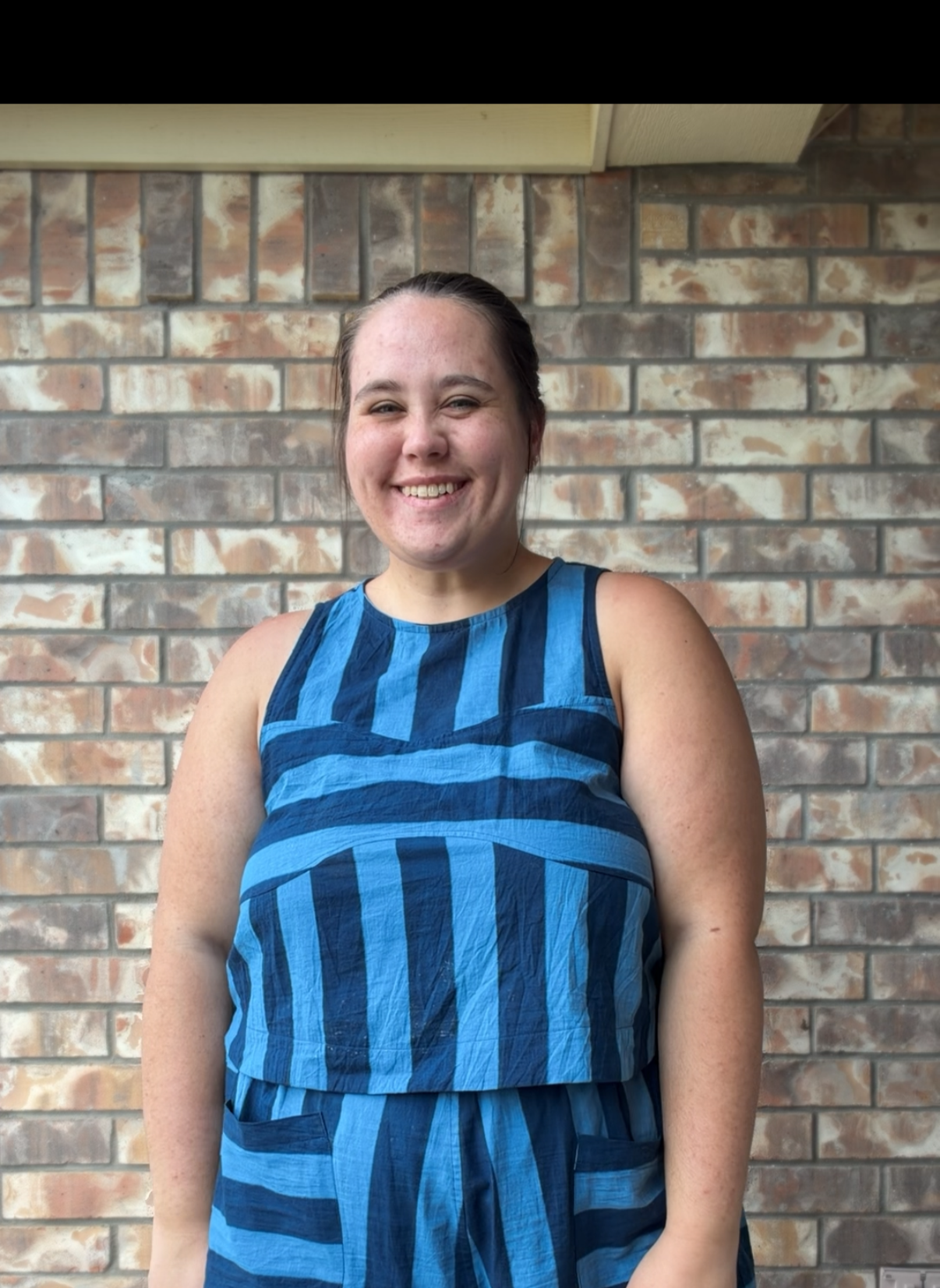 A woman smiling, standing in front of a brick wall, wearing a sleeveless blue striped top and pants set.