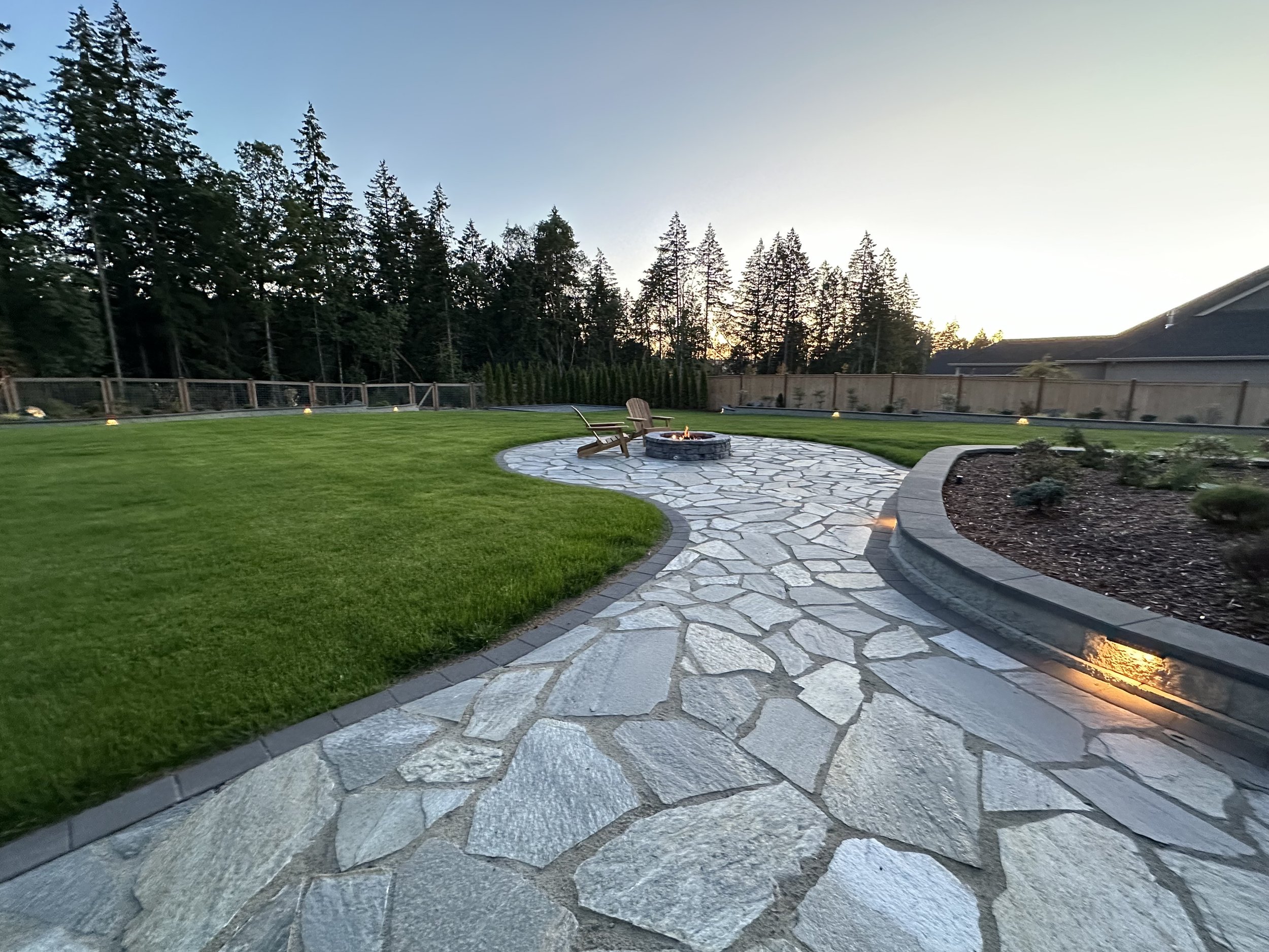 Backyard with green grass, stone patio with fire pit, two wooden chairs, surrounded by a wooden fence, trees in the background, and a clear sky at sunset.