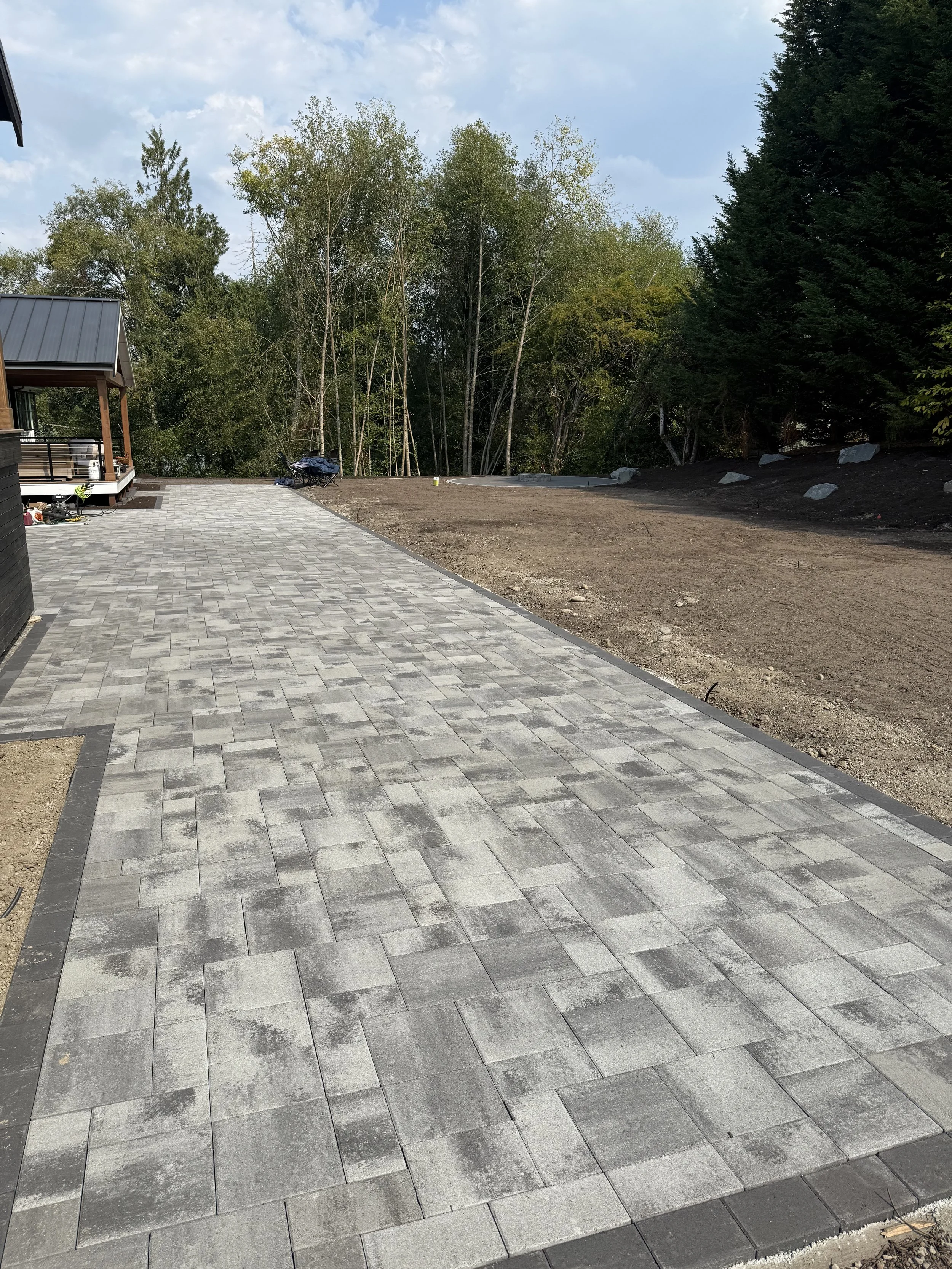 Partially completed outdoor patio with gray stone pavers and dirt area on one side, surrounded by trees and a cloudy sky.