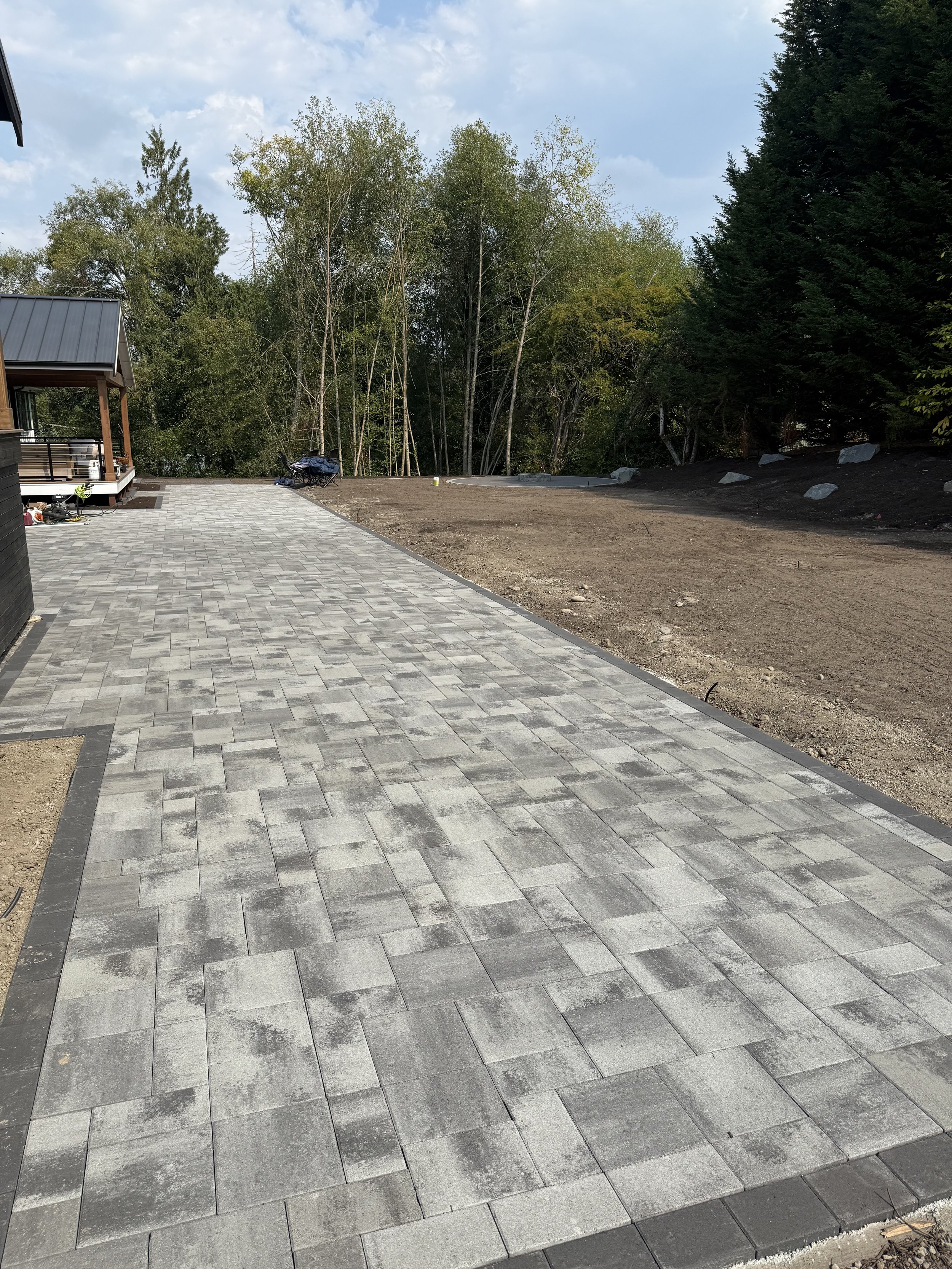 Partially completed outdoor patio with gray stone pavers and dirt area on one side, surrounded by trees and a cloudy sky.