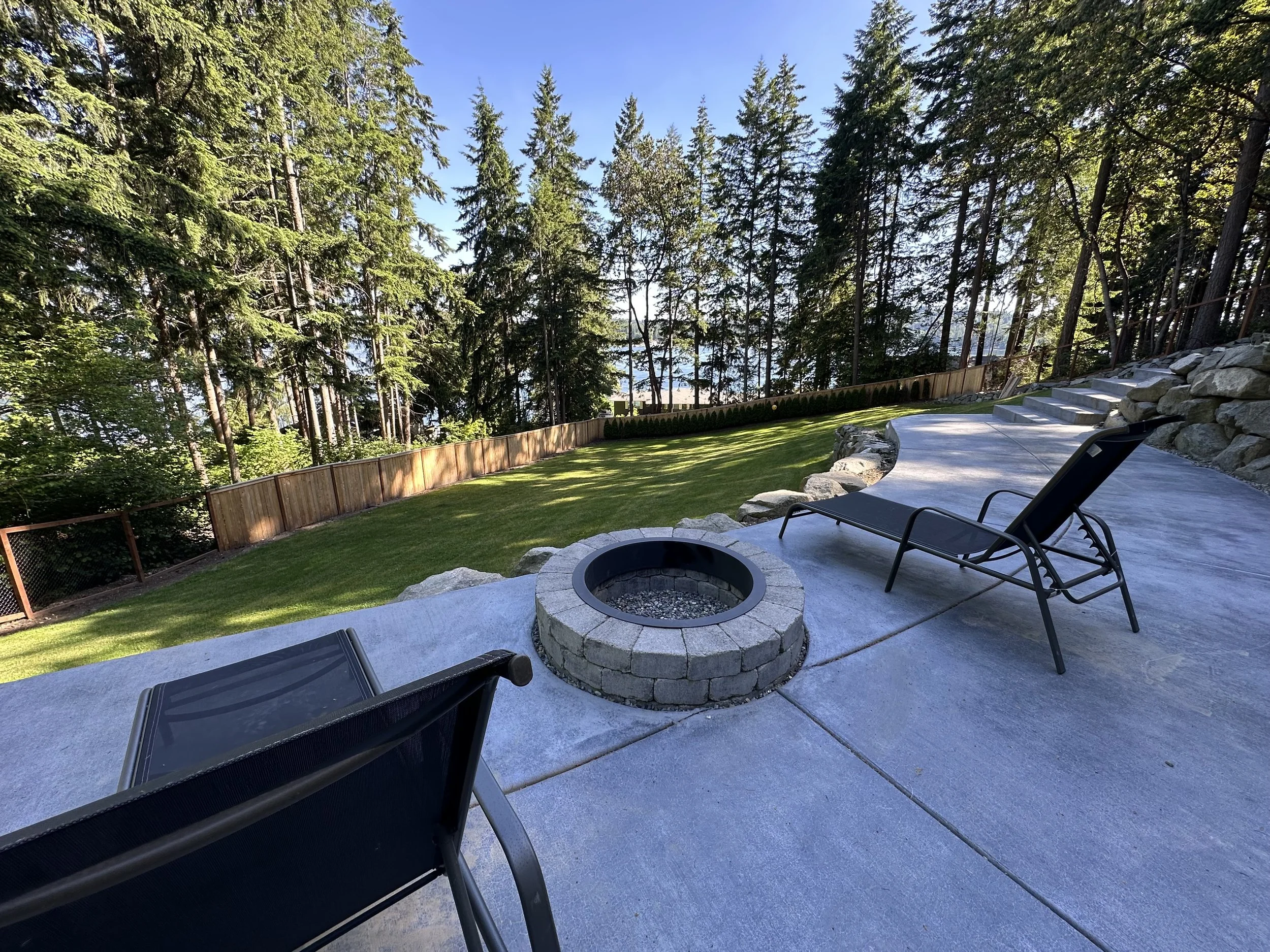A backyard patio with black lounge chairs, a fire pit, a green lawn, and tall pine trees beyond a wooden fence under a clear blue sky.