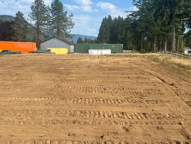A large, open dirt field with visible tire tracks, surrounded by trees and some industrial buildings in the background under a partly cloudy sky.
