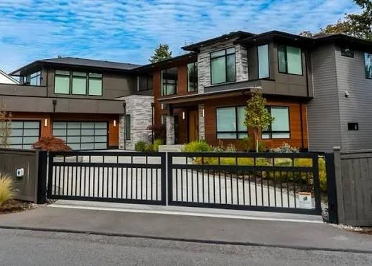 Modern two-story house with large windows, a gray and wood exterior, and a black gate at the driveway.