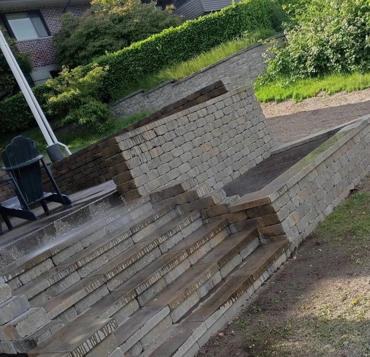 A stepped brick retaining wall with water flowing from a small fountain or water feature and a black Adirondack chair on a patio. Green bushes and grass are in the background.