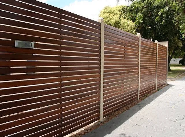 A long brown wooden privacy fence with white posts along a sidewalk, with green trees in the background.