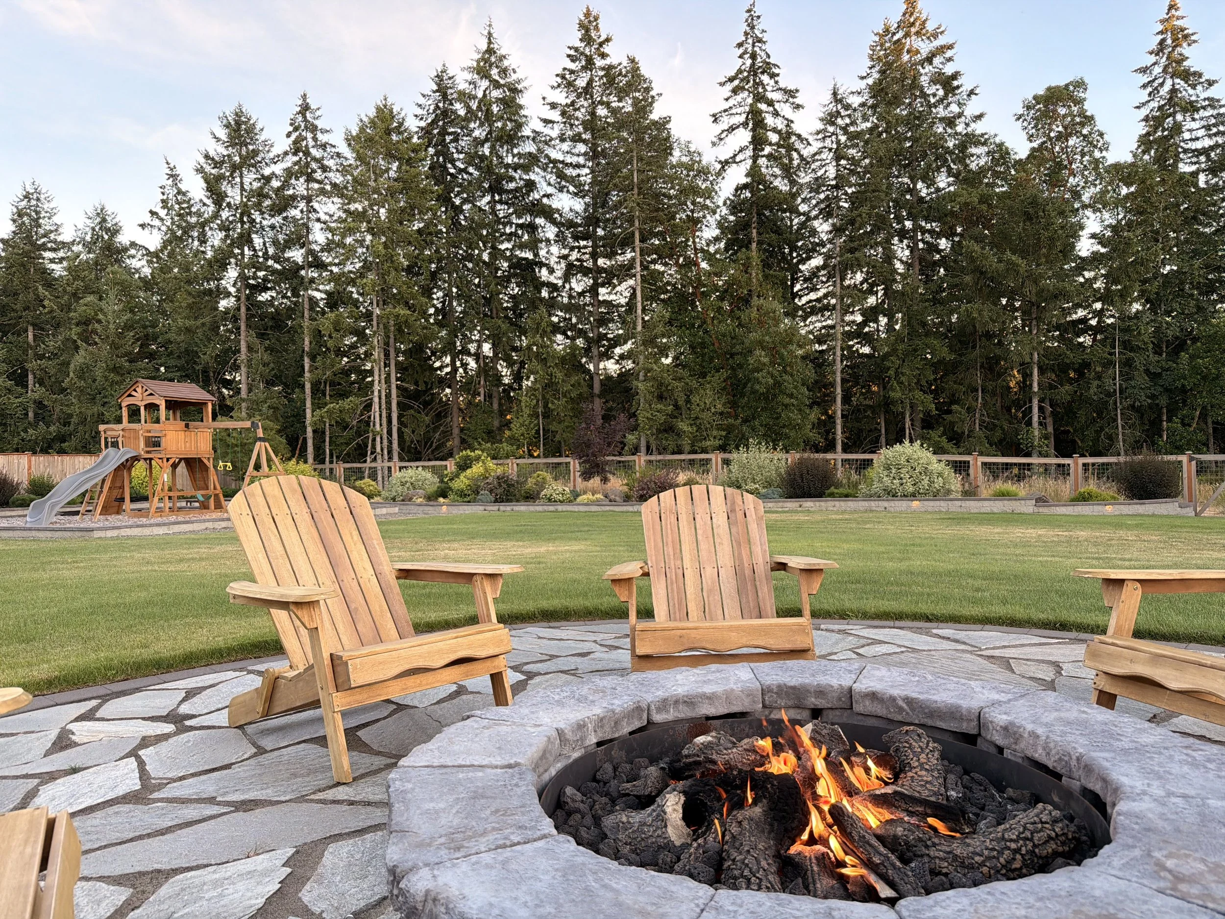 A backyard with outdoor chairs arranged around a fire pit with a small fire, a trampoline, and a children's playset with a slide. Tall trees and a fence are in the background under a partly cloudy sky.