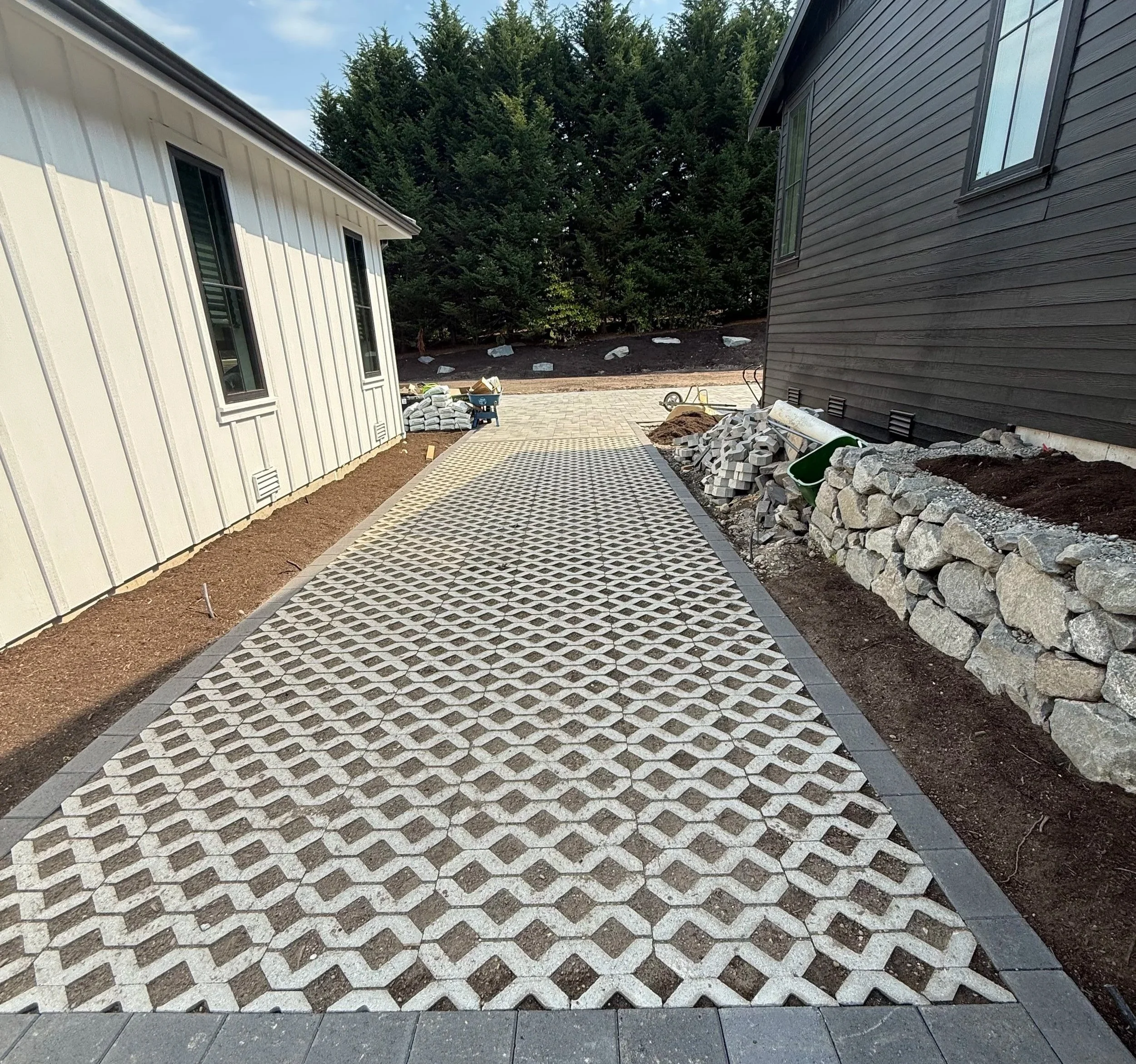Construction site with pathway of interlocking gray and white pavers, white and gray siding buildings on either side, piles of rocks, construction tools, and a backdrop of large green trees.