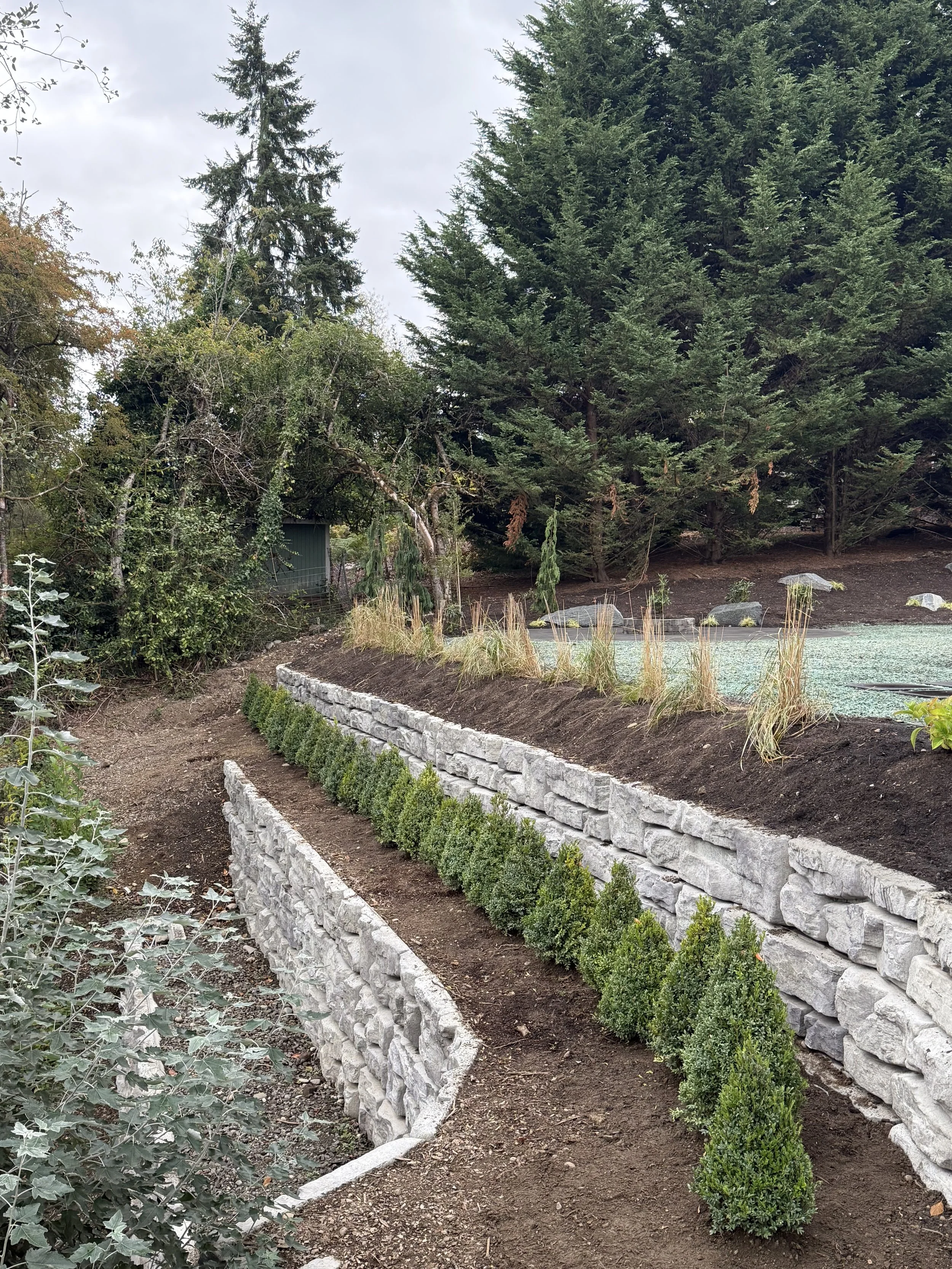 A landscaped garden with tiered stone retaining walls, neatly trimmed bushes, and a variety of trees in the background under an overcast sky.