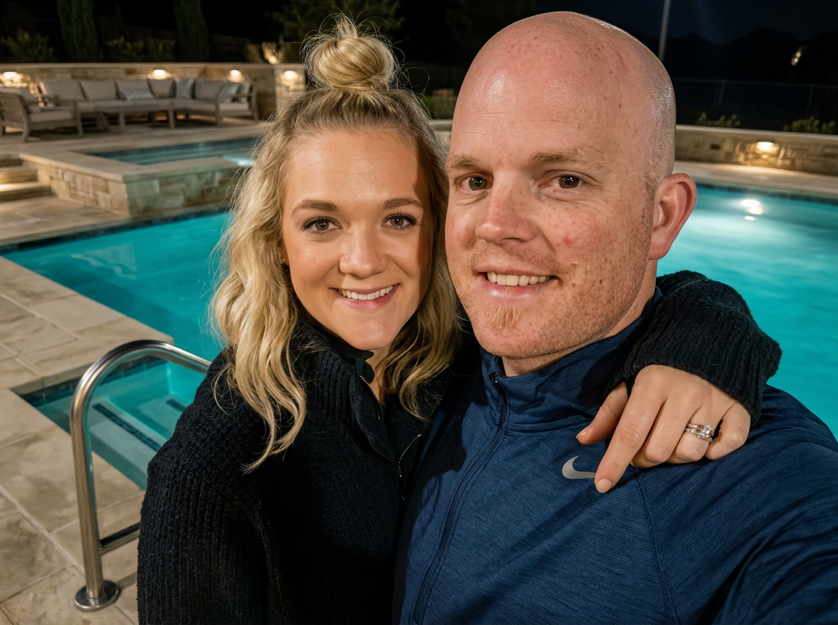 A smiling couple taking a selfie by a swimming pool at night. The woman has blonde hair styled in loose waves and is wearing a dark jacket. The man is bald, with a beard, and wearing a blue athletic jacket. The woman's hand with a wedding ring is resting on the man's shoulder.