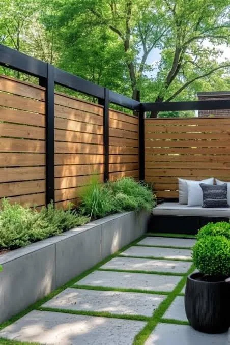 A modern outdoor backyard space with a wooden privacy fence, concrete pavers with grass in between, and potted plants, including a round shrub in a black pot, with trees and a blue sky in the background.