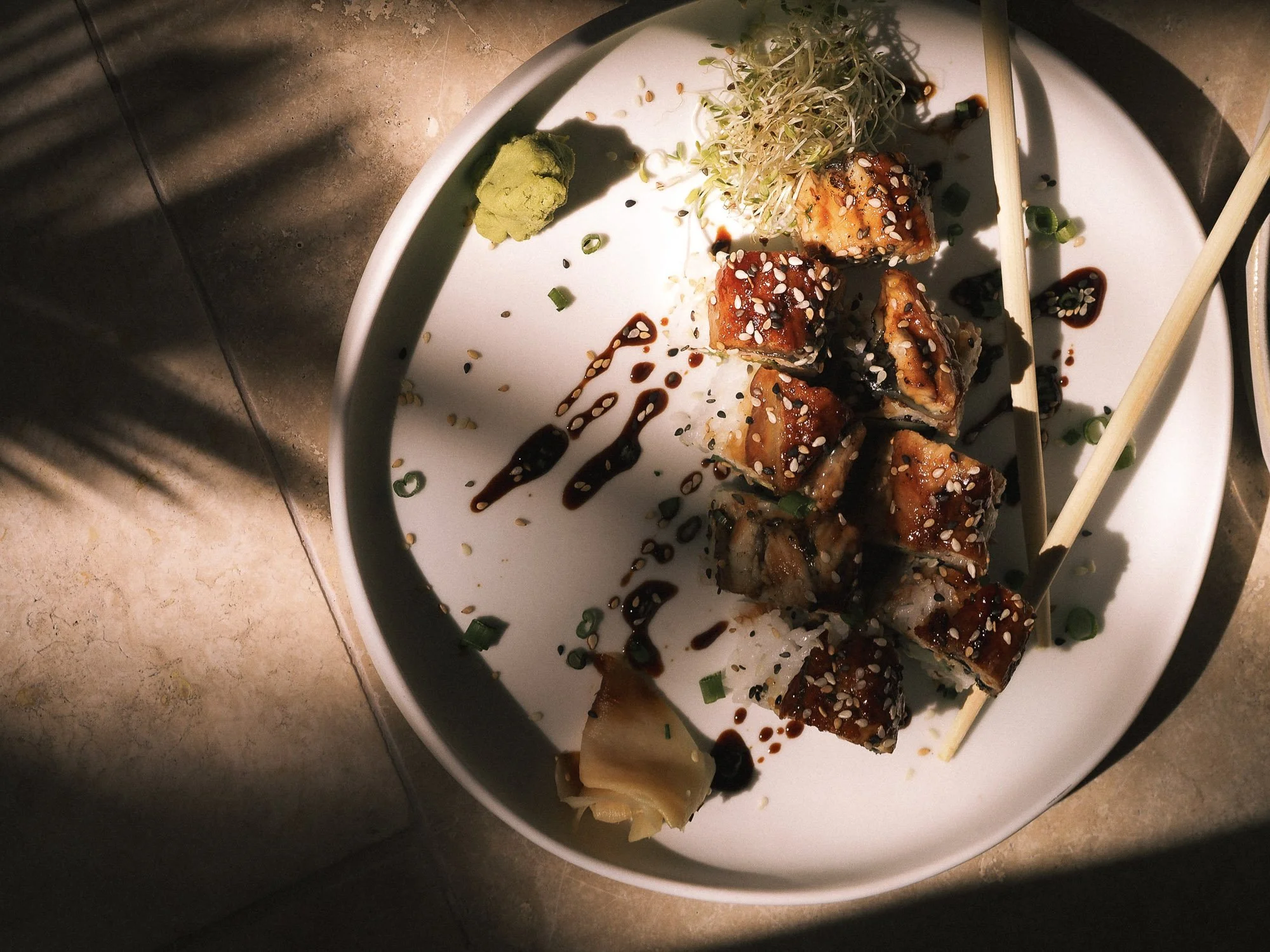 A white plate of sushi rolls drizzled with sauce, topped with sesame seeds, garnished with chopped green onions, and served with a small mound of wasabi and shredded sprouts on the side.