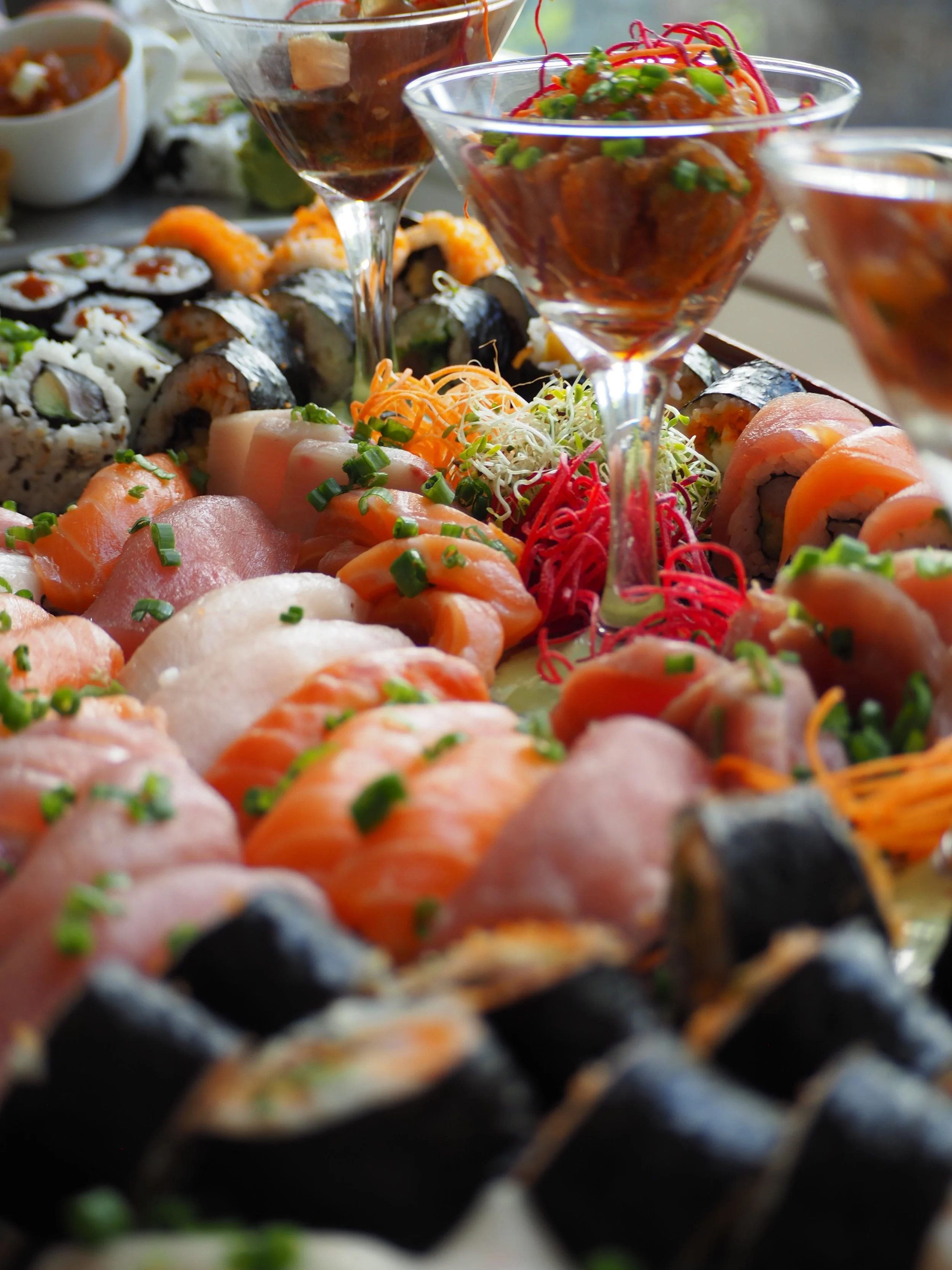 Assorted sushi rolls and sashimi on a platter with garnish, accompanied by cocktail glasses of various sauces or drinks.