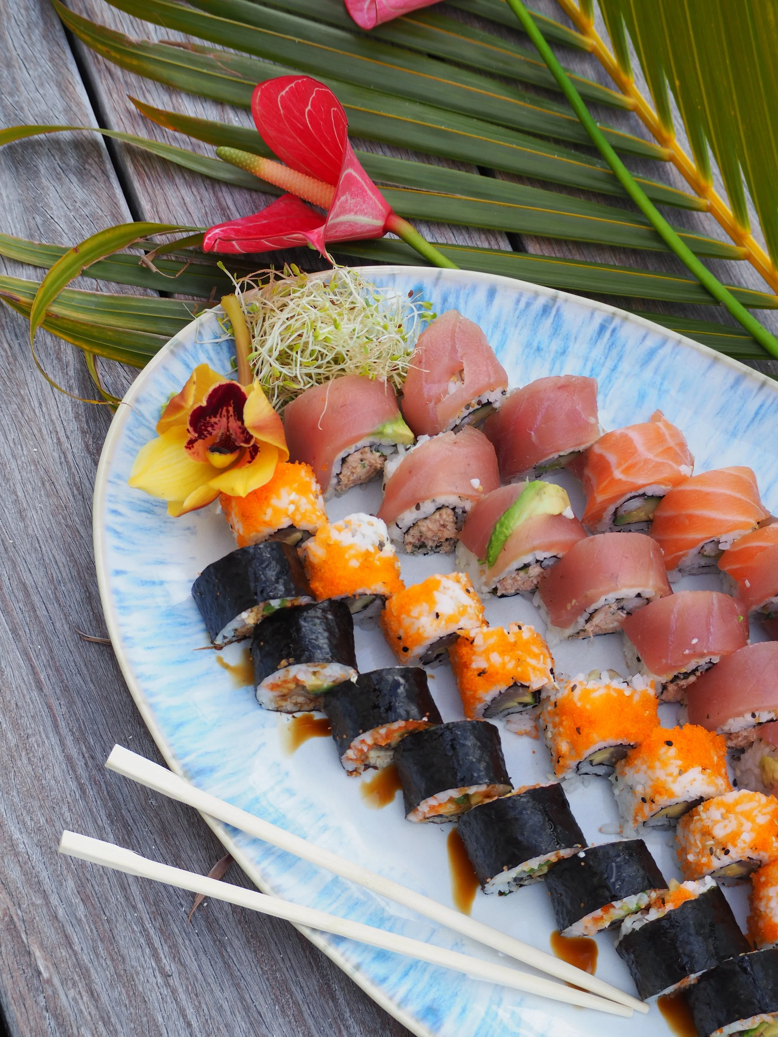 A plate of assorted sushi rolls with salmon and tuna, garnished with an orchid flower and sprouts. The plate is placed on a rustic wooden surface with tropical leaves in the background.