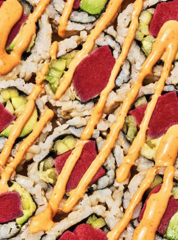 Close-up of sushi rolls topped with orange spicy mayo sauce, with cucumber, red pickled radish, and rice inside.