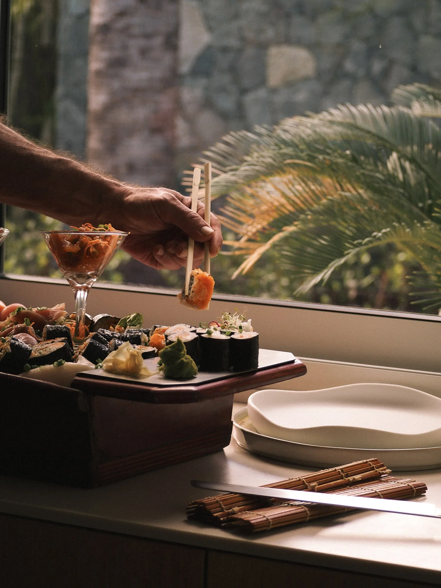 Close-up of a hand using chopsticks to pick up sushi near a window with a view of stone wall and plants outside.
