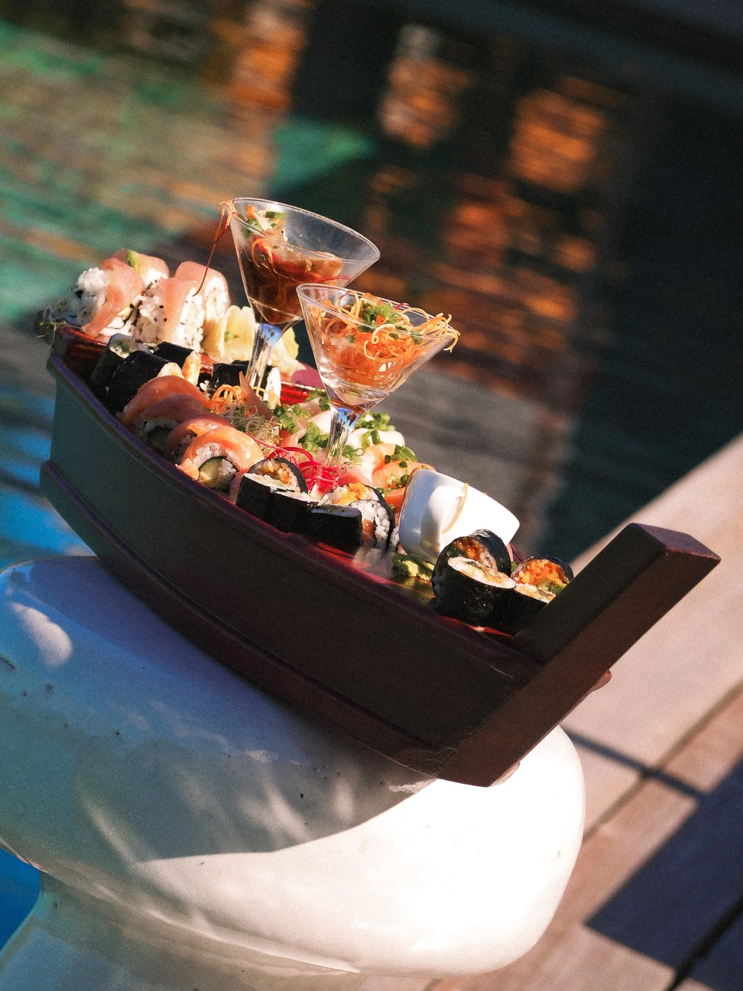 Sushi platter with various sushi rolls and two martini glasses with cocktails, served on a boat-shaped tray.