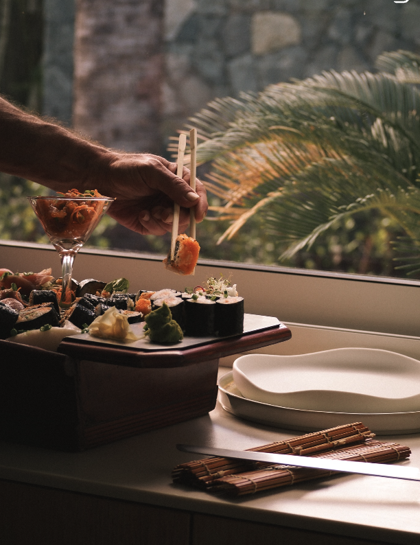 Person using chopsticks to pick up sushi from a wooden platter on a windowsill, with a martini glass and empty plates nearby, with palm leaves visible outside.