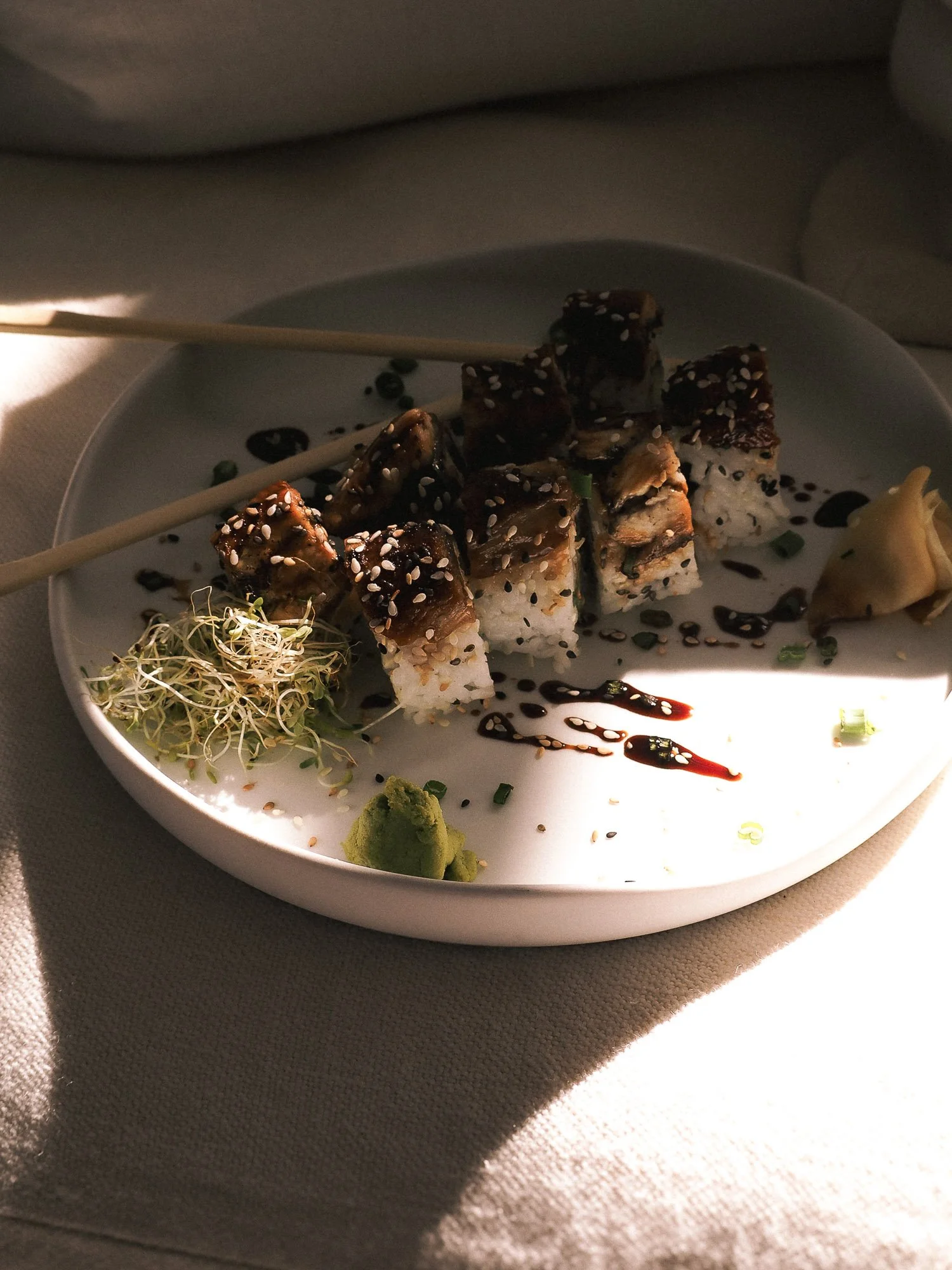 Sushi rolls topped with grilled eel and sesame seeds, served with wasabi, ginger, soy sauce, and microgreens, on a white plate.