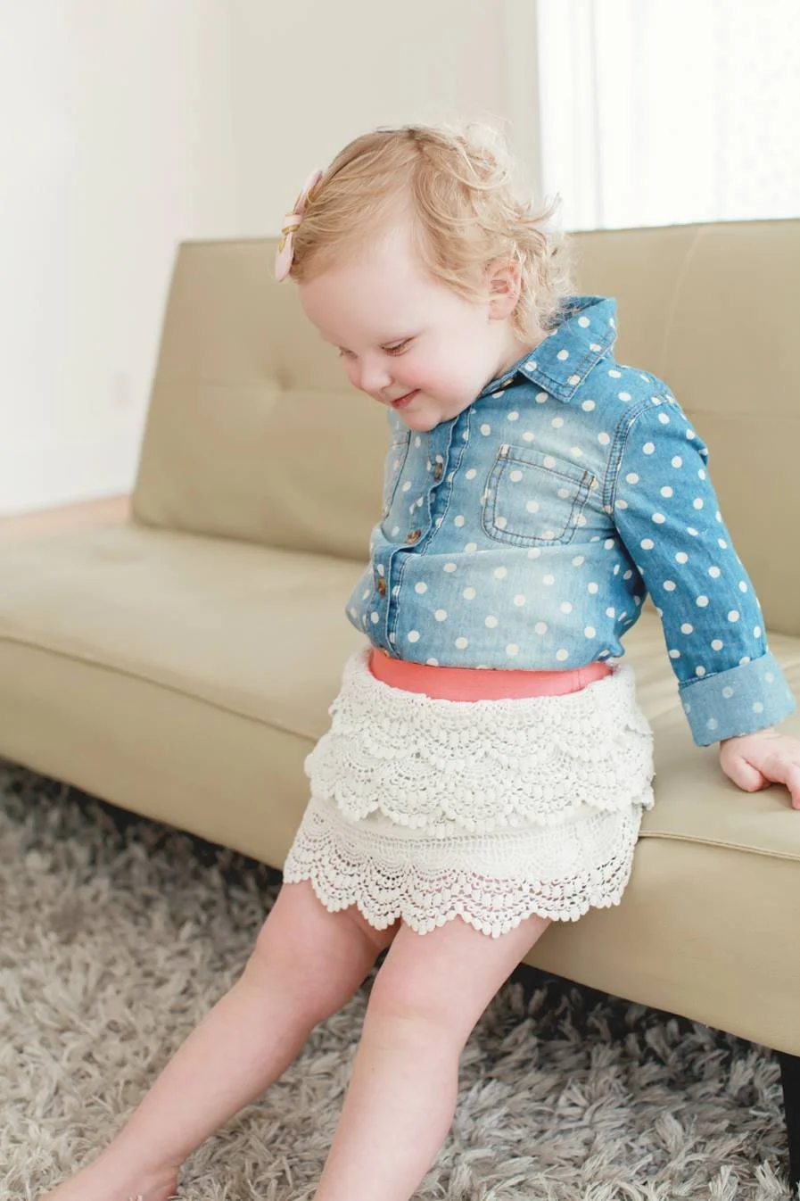 A young girl with blonde curly hair, wearing a pink hair bow, a blue polka dot denim shirt, a peach top, and a white lace skirt, sitting on a beige sofa in a bright room.