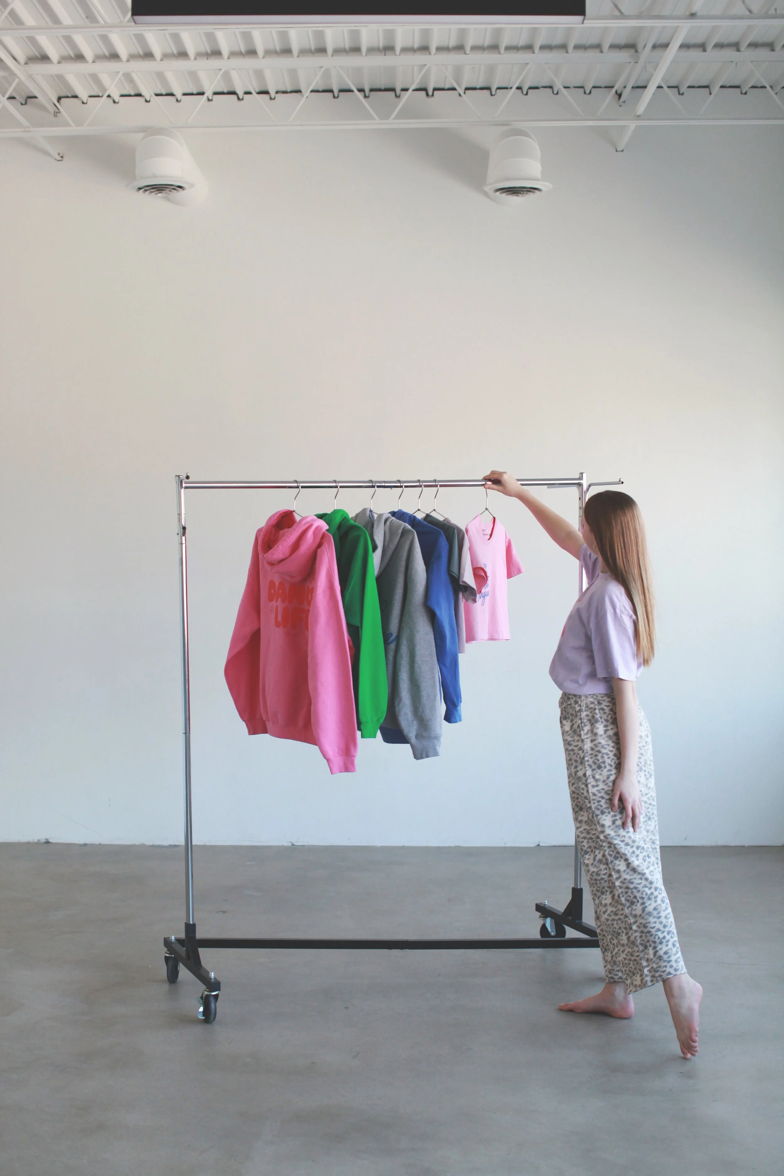 A woman with long hair and patterned pajama pants touching a clothing rack with various colored hoodies and T-shirts in a room with a high ceiling and exposed beams.