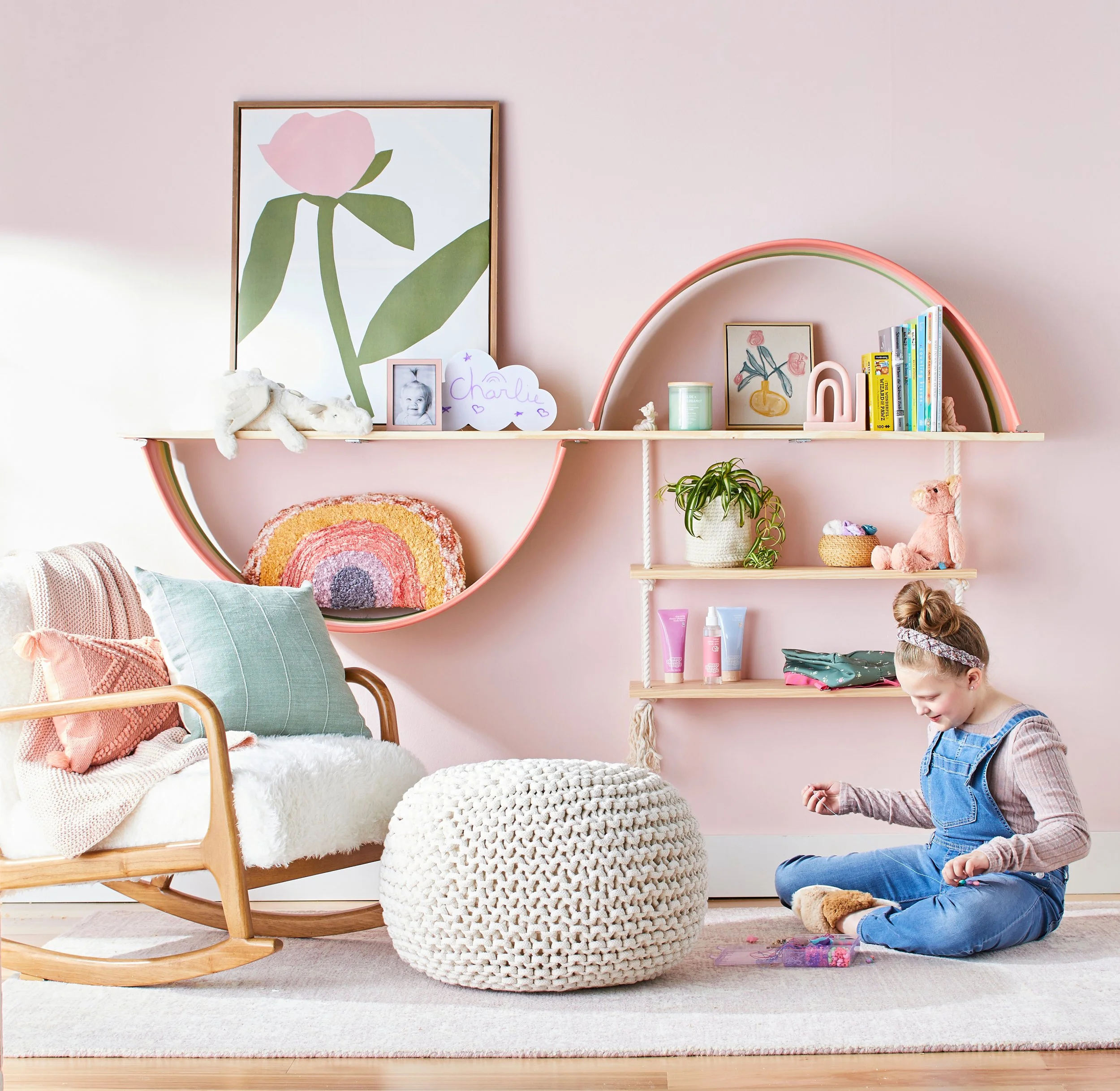 A pink wall-mounted shelf with a rainbow-shaped frame. On the shelf are a potted plant, a framed artwork of flowers in a vase, a small framed photograph of a child's face, a white cloud-shaped sign with purple writing that says 'Charlie' with purple hearts, a mint green candle, a pink sculpture, a stack of children’s books, and some tubes of skincare or beauty products. In front of the shelf are a knitted pouf, a tufted fluffy white blanket on a rattan rocking chair, and a young girl sitting on the floor, playing with beads or jewelry, wearing a denim jumper and a headband.
