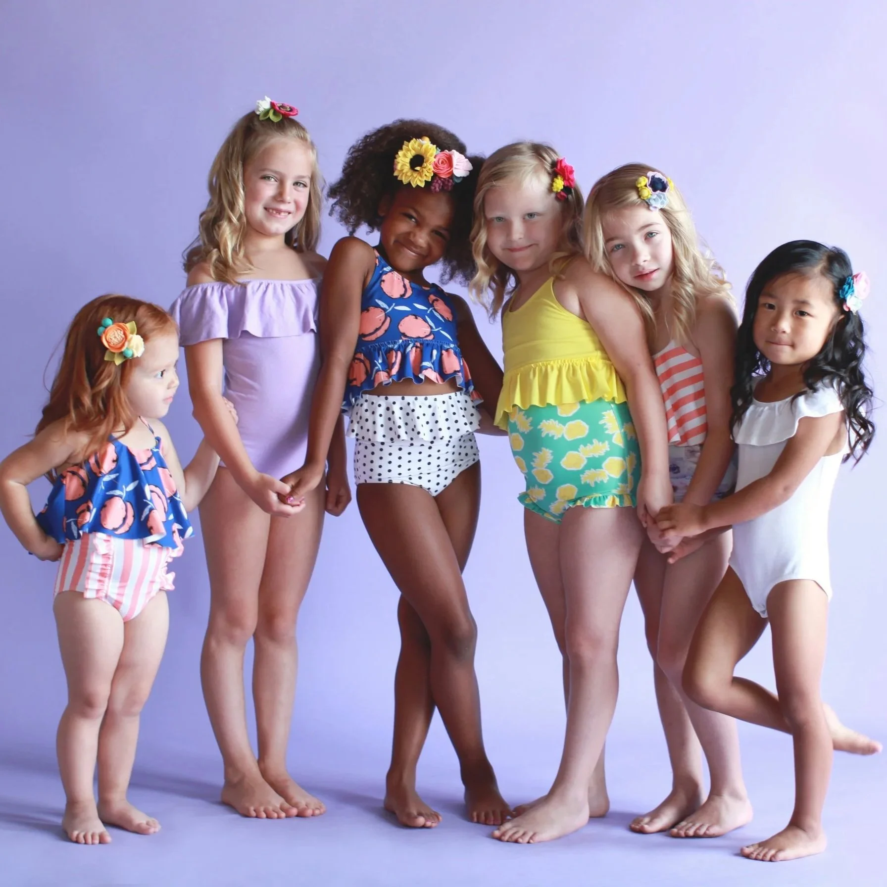 Group of young girls with colorful flowers in their hair, wearing summer dresses and shorts, standing together against a purple background.
