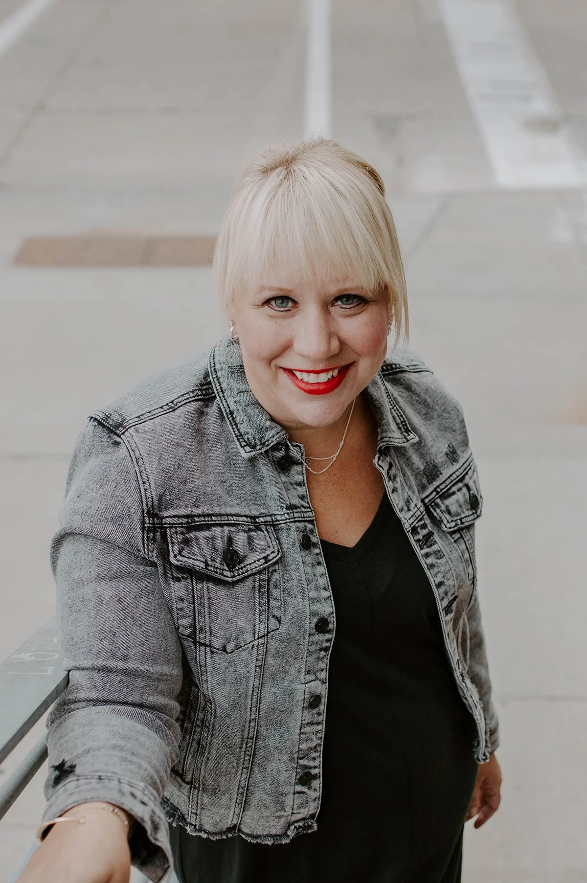 A smiling woman with platinum blonde hair, red lipstick, and blue eyes wearing a gray denim jacket over a black top, sitting outdoors on a concrete surface.