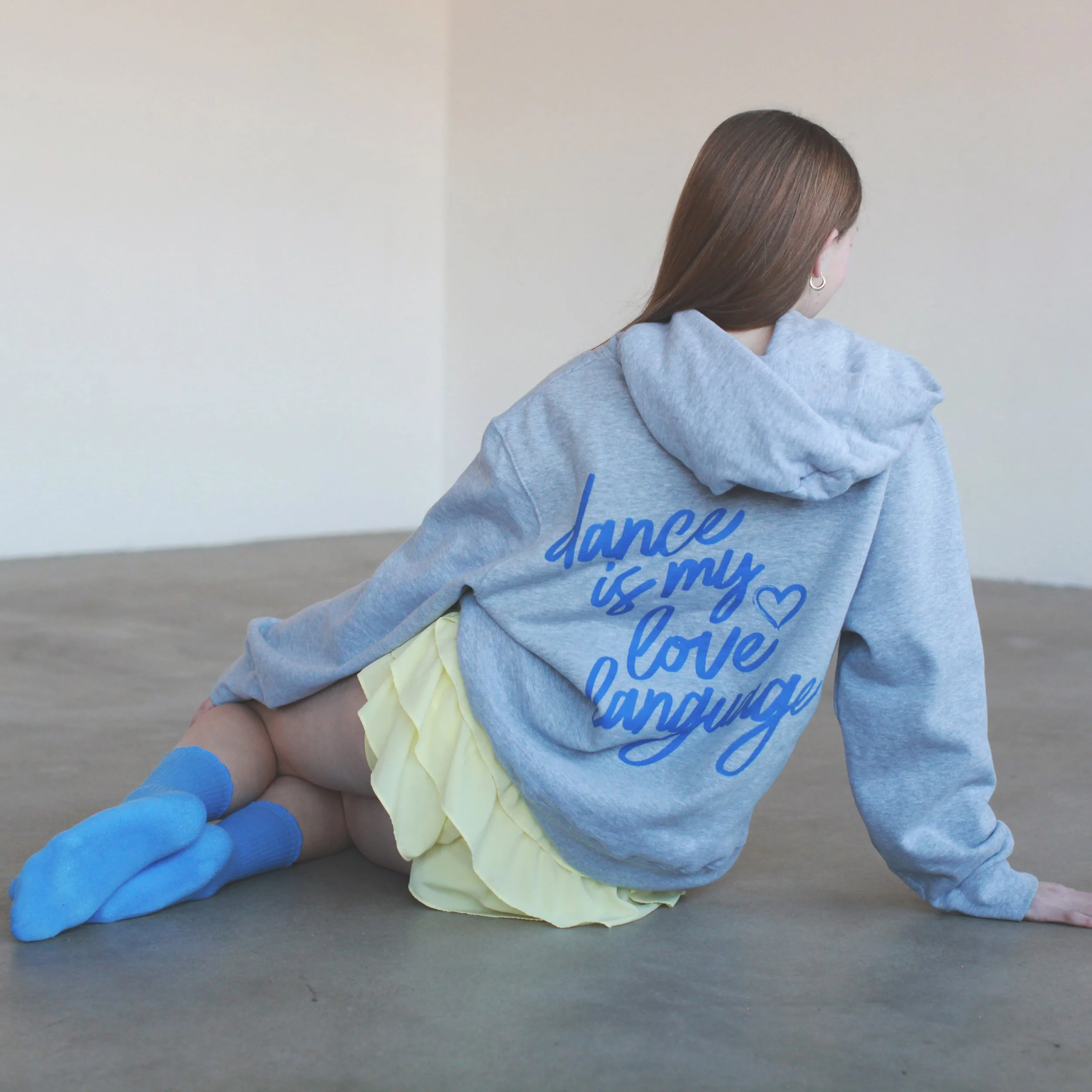 A person wearing a gray hoodie is lying on the ground with their back to the camera, wearing yellow shorts and blue gloves. The person has brown hair and is sitting against a plain wall.