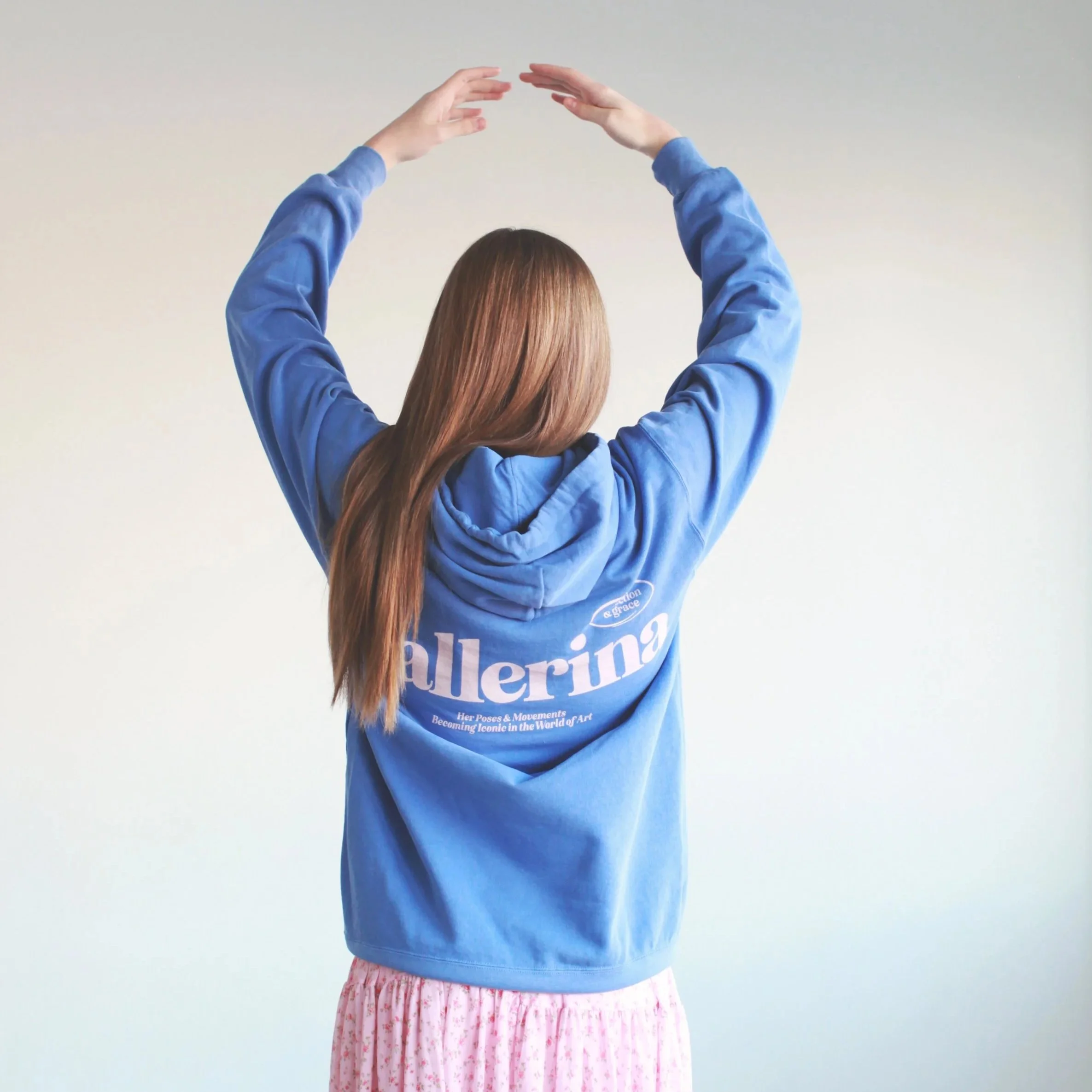 Person with long hair wearing a blue sweatshirt and pink floral skirt, standing against a plain background, arms raised with hands forming a shape above their head.