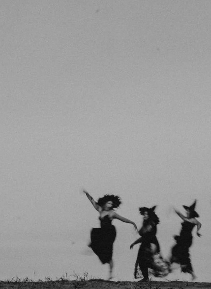 Three women dancing on a grassy hilltop, wearing wide-brimmed hats and flowing dresses, with a clear sky in the background.
