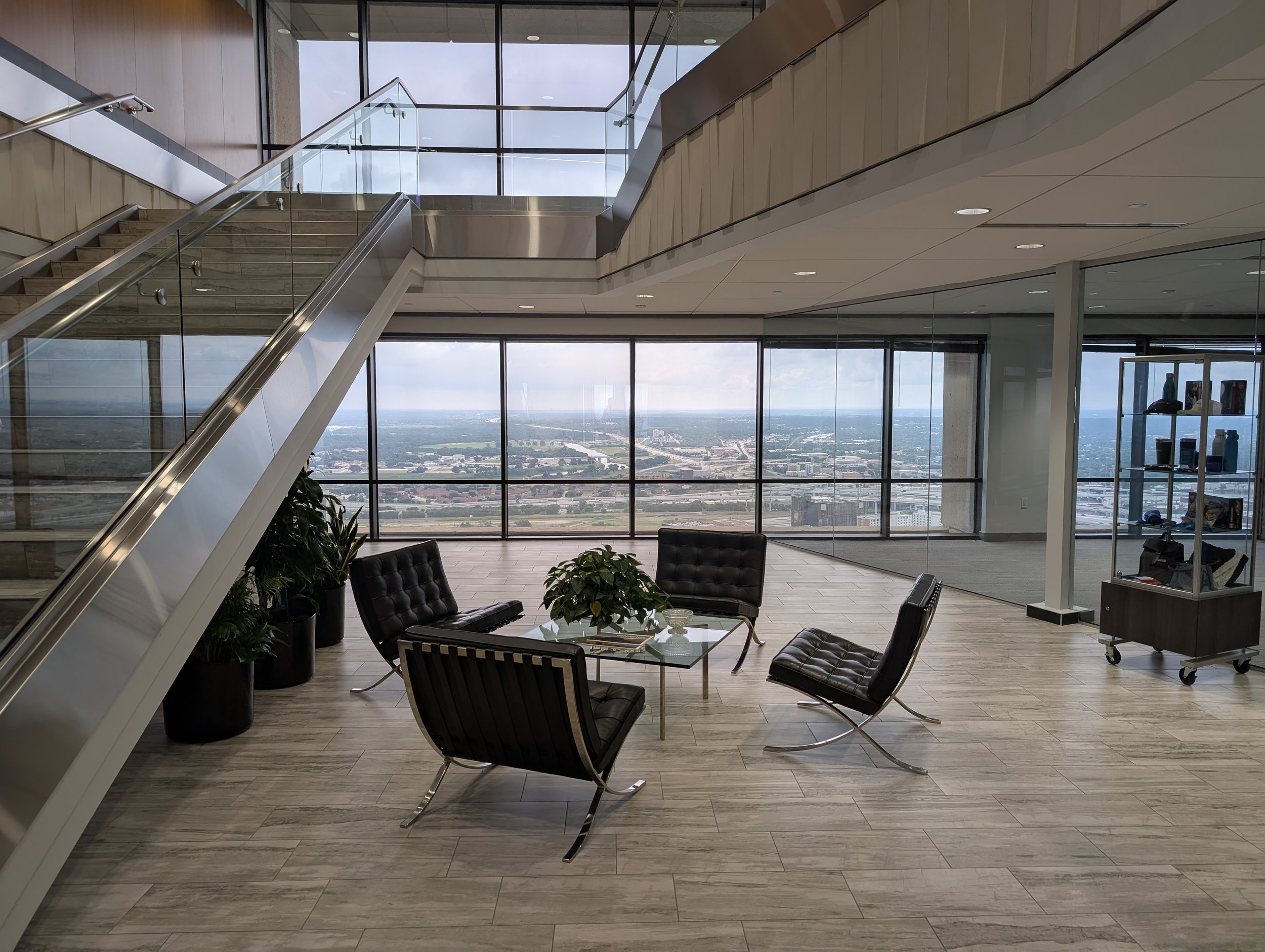 Interior of a modern office or waiting area with large floor-to-ceiling windows showing an expansive city view. The space contains black leather chairs, round glass table, and potted plants. A staircase with glass railing leads to an upper level, and there are ceiling lights. A display shelf with various items is visible on the right.