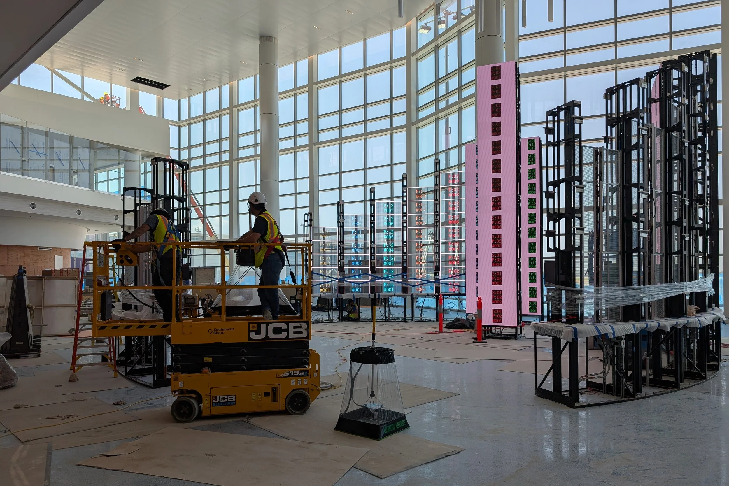 Interior of a modern building under construction, with large glass windows, construction workers, industrial equipment, scaffolding, and digital display boards.