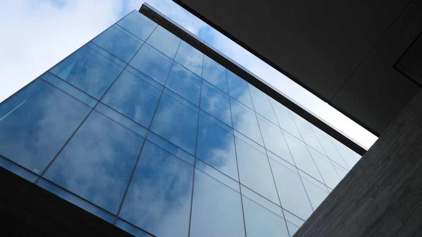 Looking up at a modern glass building with blue sky and clouds reflected on its surface, featuring sharp angles and a minimalist design.