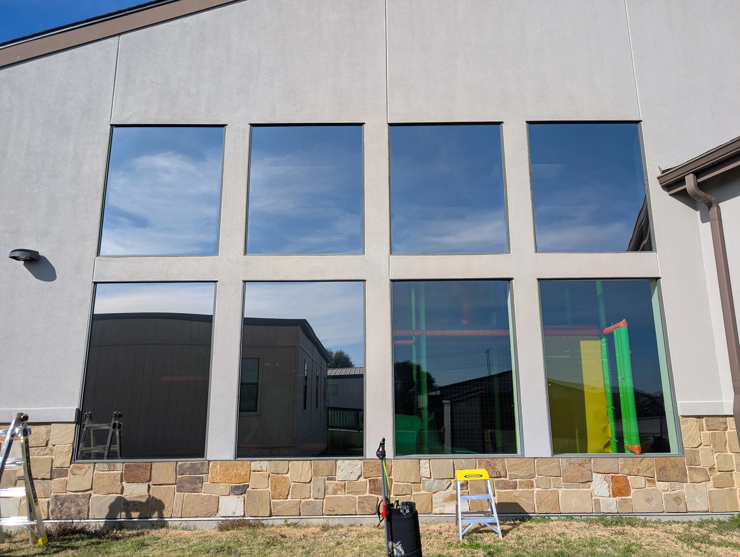 The image shows a modern building with large rectangular windows reflecting the sky and clouds. There are some construction or cleaning tools and ladders at the bottom of the building, indicating ongoing work. The building has a stone foundation and a light-colored textured exterior wall.