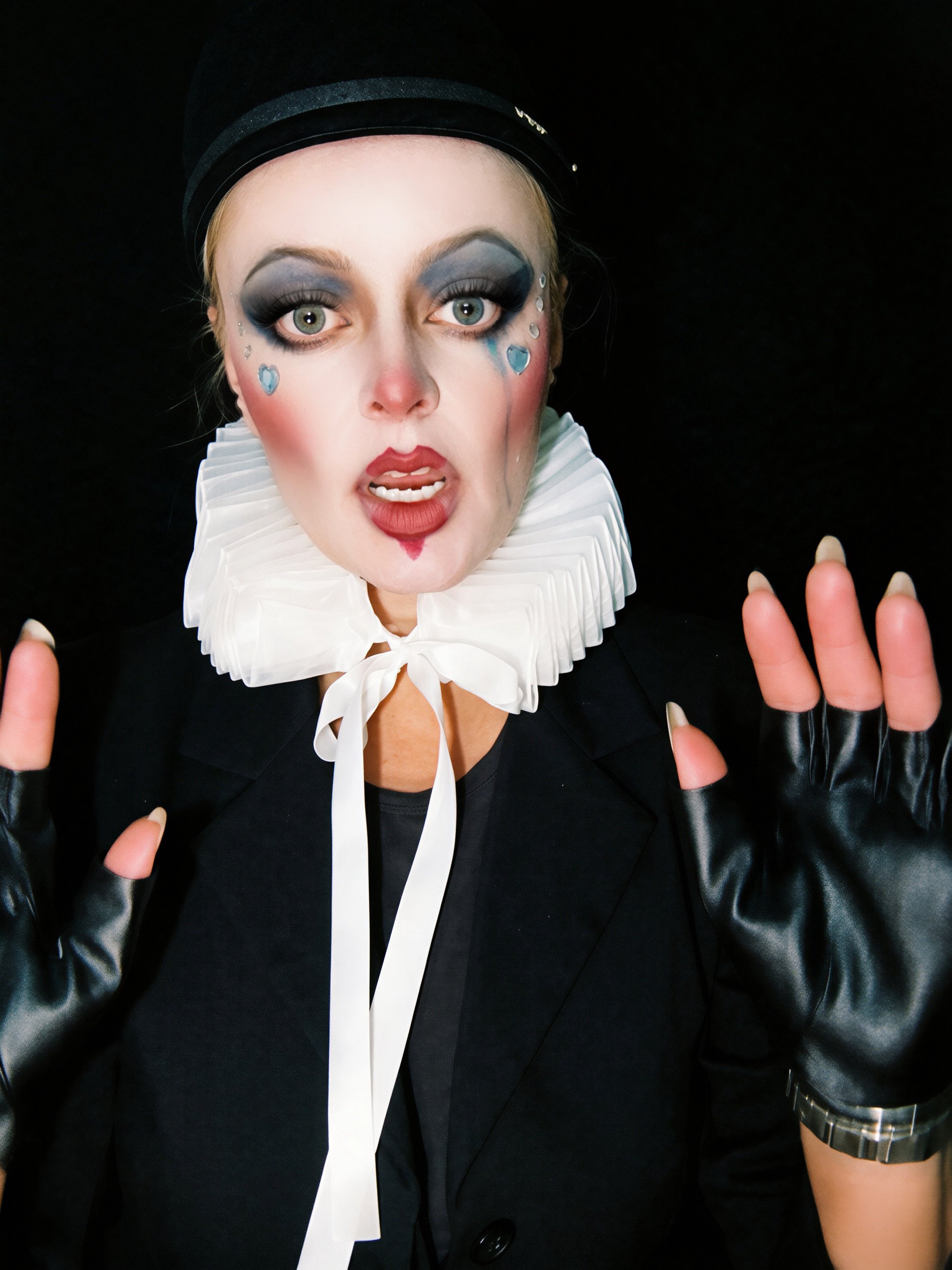 Person with dramatic makeup, gray eyeshadow, black eyeliner, and face rhinestones, wearing a black hat, ruffled white collar, black jacket, and black gloves, posing with hands near face against a black background.