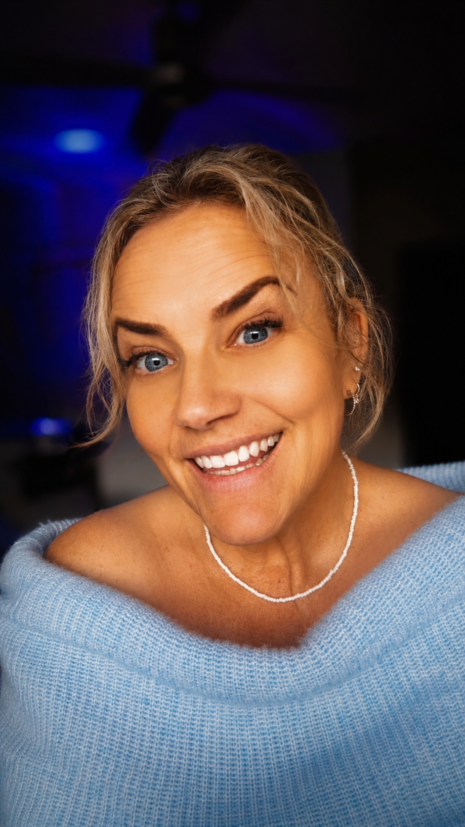 Close-up portrait of a smiling woman with blue eyes, short blonde hair, wearing a light blue sweater, pearl necklace, and earrings, with dark background and blue lighting.