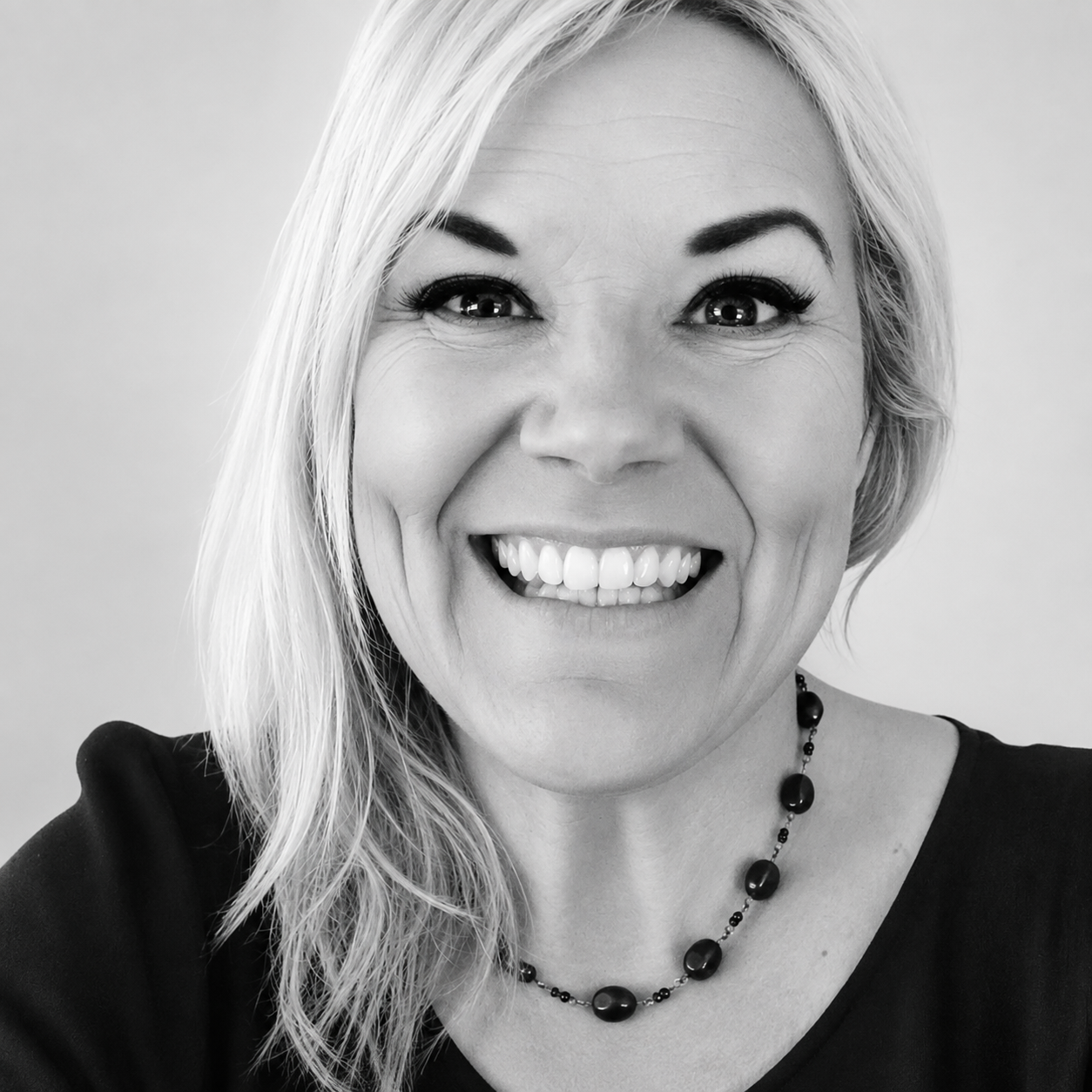 Close-up black-and-white portrait of a smiling woman with light-colored hair, wearing a dark top and a beaded necklace.