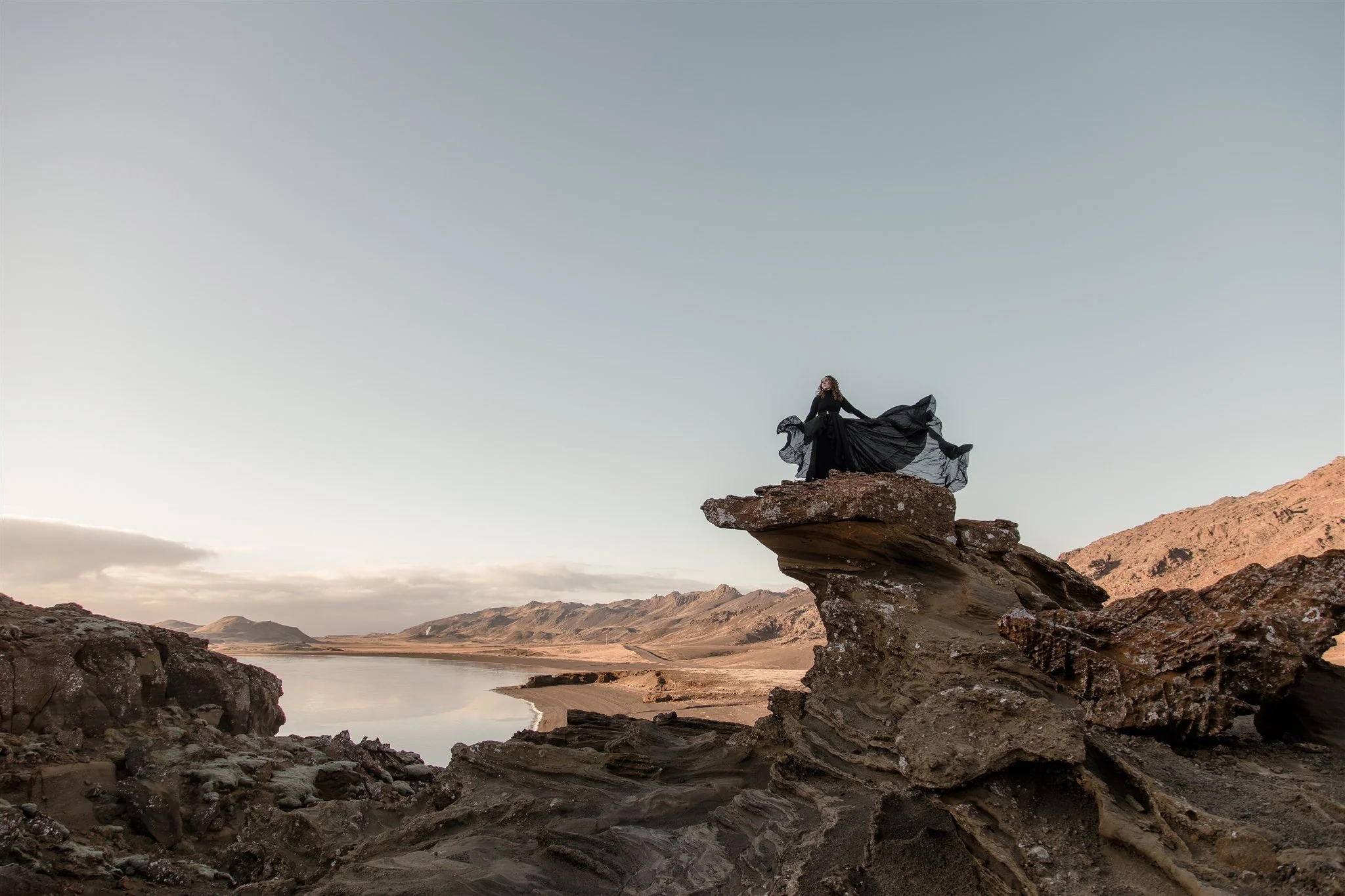 A woman in a black dress with a flowing skirt is sitting on a large rock formation overlooking a landscape with rugged mountains, a valley, and a river, under a cloudy sky.