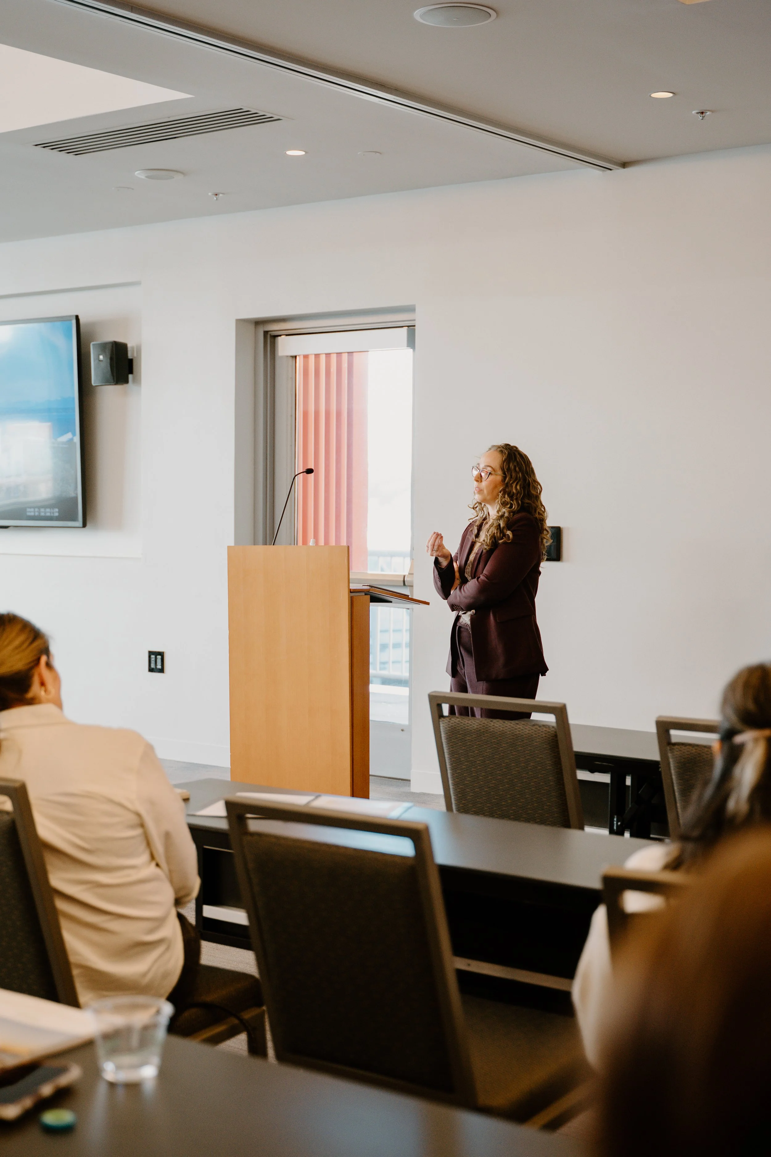 A woman in a maroon blazer and glasses is speaking at a podium in a conference room with several seated attendees, large window with blinds, wall-mounted TV, and ceiling lights.